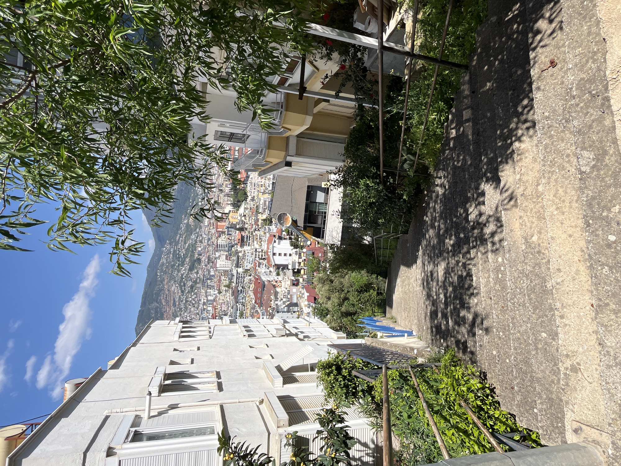 The image depicts a view looking down a set of concrete steps, framed by buildings and vegetation, with a cityscape in the distance. The steps lead downward and right, with a metal railing on the left side. A variety of plants and trees are visible along the steps and in the foreground, casting shadows on the concrete.
On the left, a white building with multiple windows is partially visible. Further in the distance, a densely built-up city stretches out, backed by a mountain under a blue sky with some clouds. The city is Alanya, Turkey, identifiable by the urban sprawl climbing the hillside and the architecture.