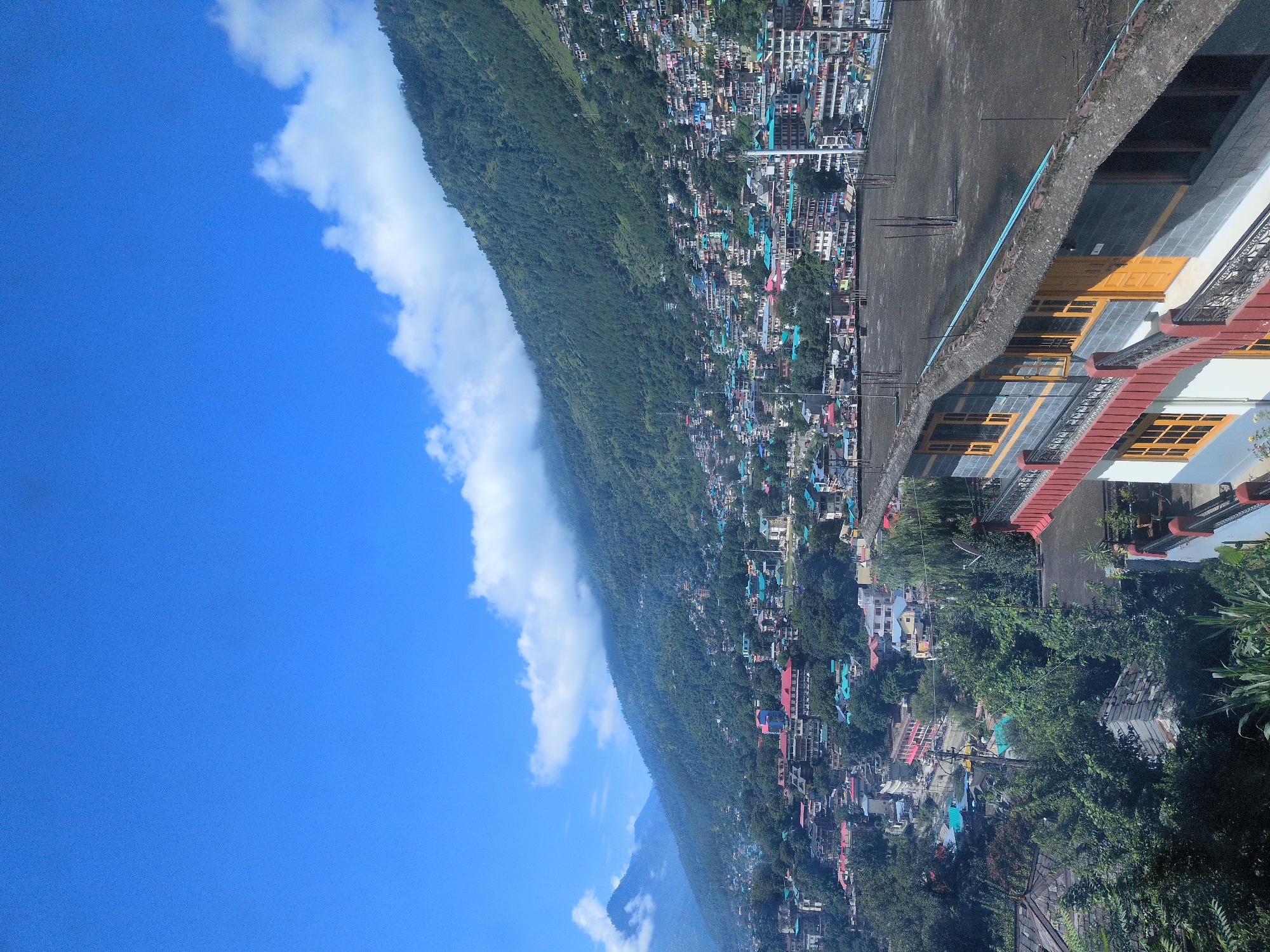 A high-angle shot captures the bustling city of Kullu, India, nestled amidst lush green mountains under a vibrant blue sky dotted with white clouds. The city, a dense tapestry of colorful buildings with red, blue, and teal roofs, sprawls across the steep slopes of the mountains. Narrow streets wind through the urban landscape, barely visible between the tightly packed structures. In the foreground, the roof of a dark-tiled building with a red and white facade is prominent. This building features yellow-framed windows and wooden doors, hinting at traditional architecture. The mountainside is densely forested, with trees reaching up to where the buildings begin. The sky is clear and bright, suggesting a sunny day. The overall impression is one of a densely populated town integrated into a dramatic natural environment.