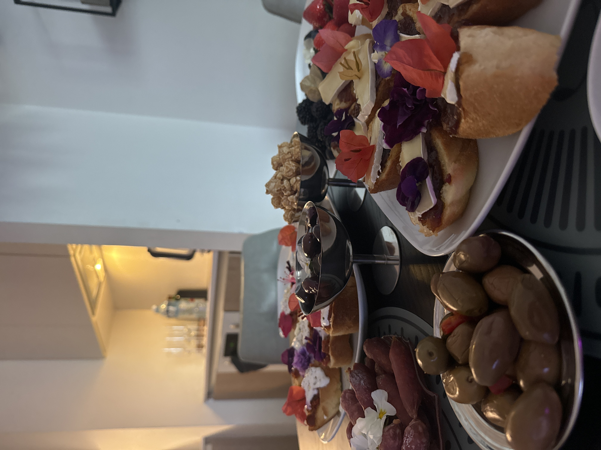 The image presents a close-up of a tiered serving platter laden with various food items. The scene likely depicts a buffet or appetizer spread at a gathering. The food items include: baguette slices with what appears to be brie cheese and toppings, possibly including jam or compote and garnished with edible flowers. There are also bowls containing popcorn, olives, and what seems to be salami. The platter is arranged in a tiered fashion, suggesting an attempt at visual presentation. The location is not evident from the food or tableware alone. The overall impression is that of a casual yet carefully arranged food display.