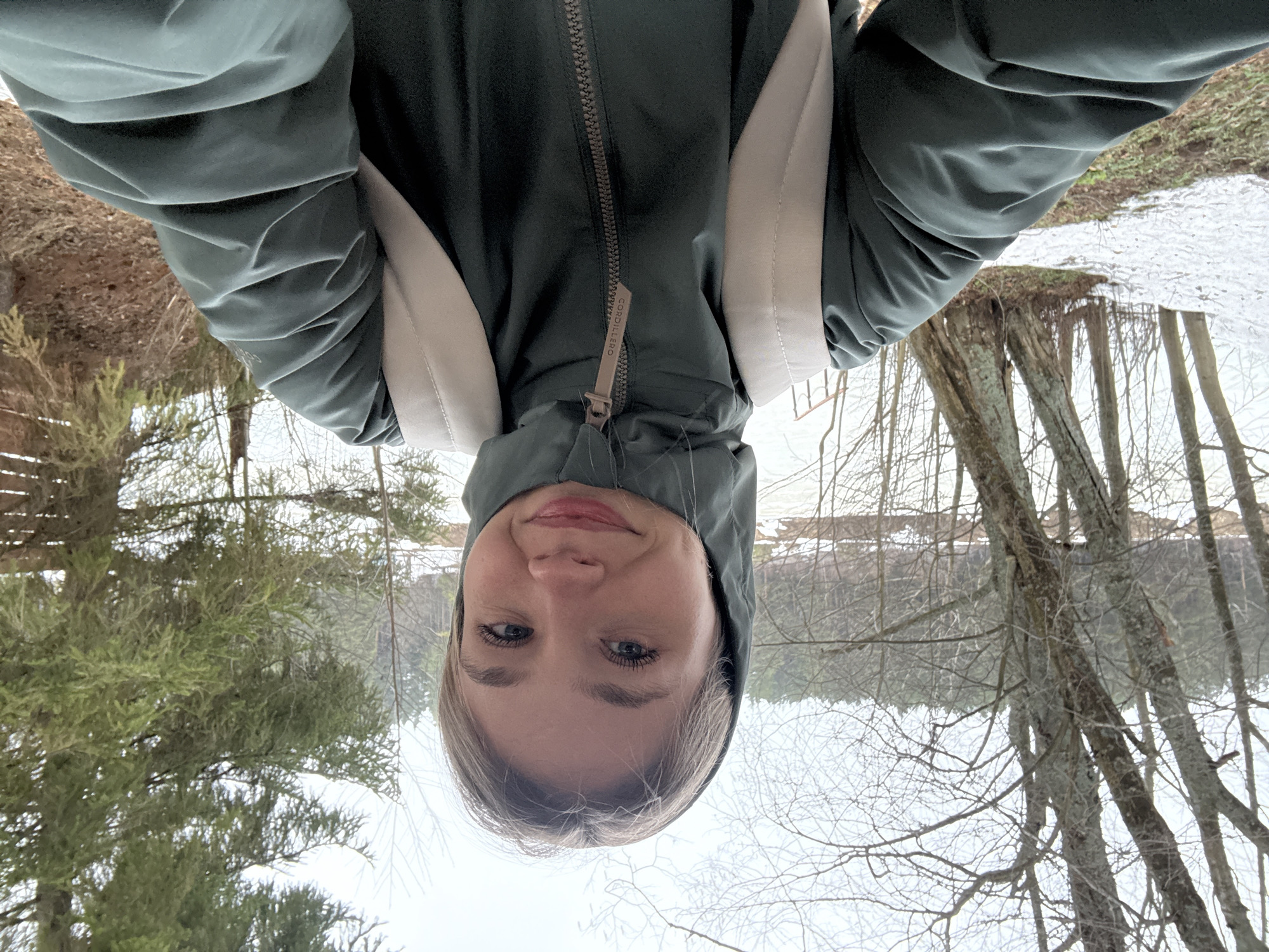 A selfie perspective shows a fair-skinned female wearing a hooded green jacket with a zipper and beige accents, seemingly in an outdoor environment. The background features bare trees, some with snow near the base, and possibly a body of water reflecting the scene. The location appears to be a natural area or park, though precise geolocation within Chaikovsky, Russia cannot be determined from the image. No specific actions or interactions are discernible.