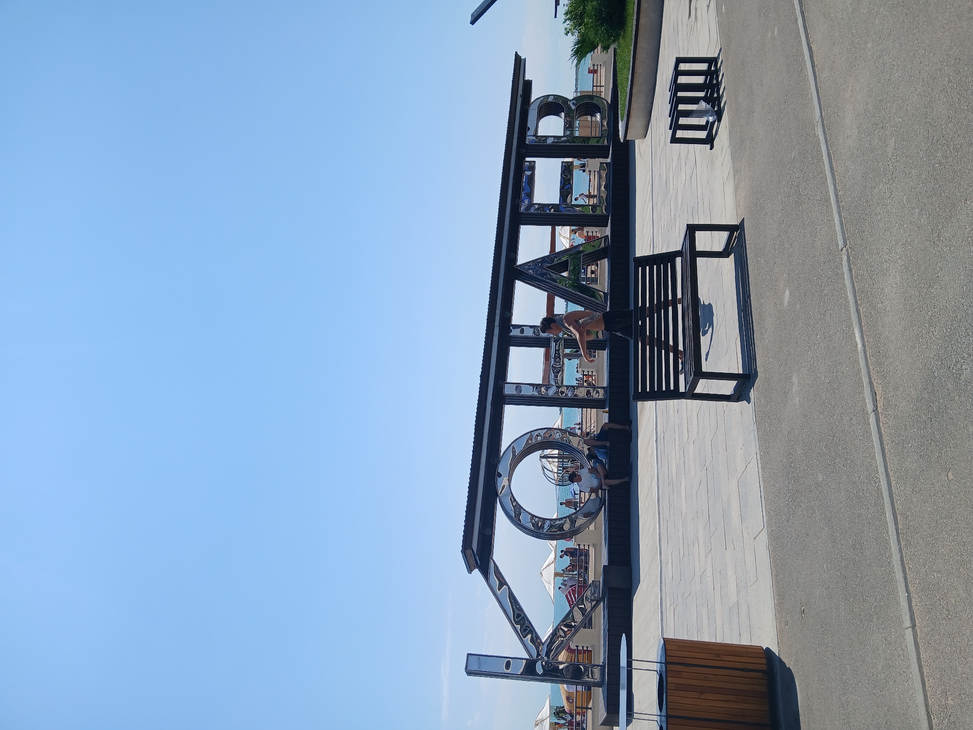 In the city of Qonaev, Kazakhstan, on a bright, clear day, a large, modern sculpture spelling out "KOНАЕВ" in reflective metal letters stands prominently. The sculpture is positioned in front of a body of water, likely the Kapchagay Reservoir, with a beach area visible in the background. Several white beach umbrellas are dotted along the shore.
In the foreground, a paved plaza with a clean, geometric design surrounds the sculpture. A dark wooden trash receptacle with metal accents is situated to the left of the plaza. A black metal bench is placed in the center of the plaza, with a person walking past it. To the right of the bench, a small, black metal rack, possibly for bicycles, is visible.
Several people are present in the scene. Two individuals are seated on a dark platform in front of the sculpture. In the background, on the beach, more people are seen relaxing and walking. One person, a young man, is captured mid-stride on the plaza, wearing dark shorts and carrying a towel over his shoulder. The overall atmosphere is one of a relaxed, sunny day at a popular public space.
