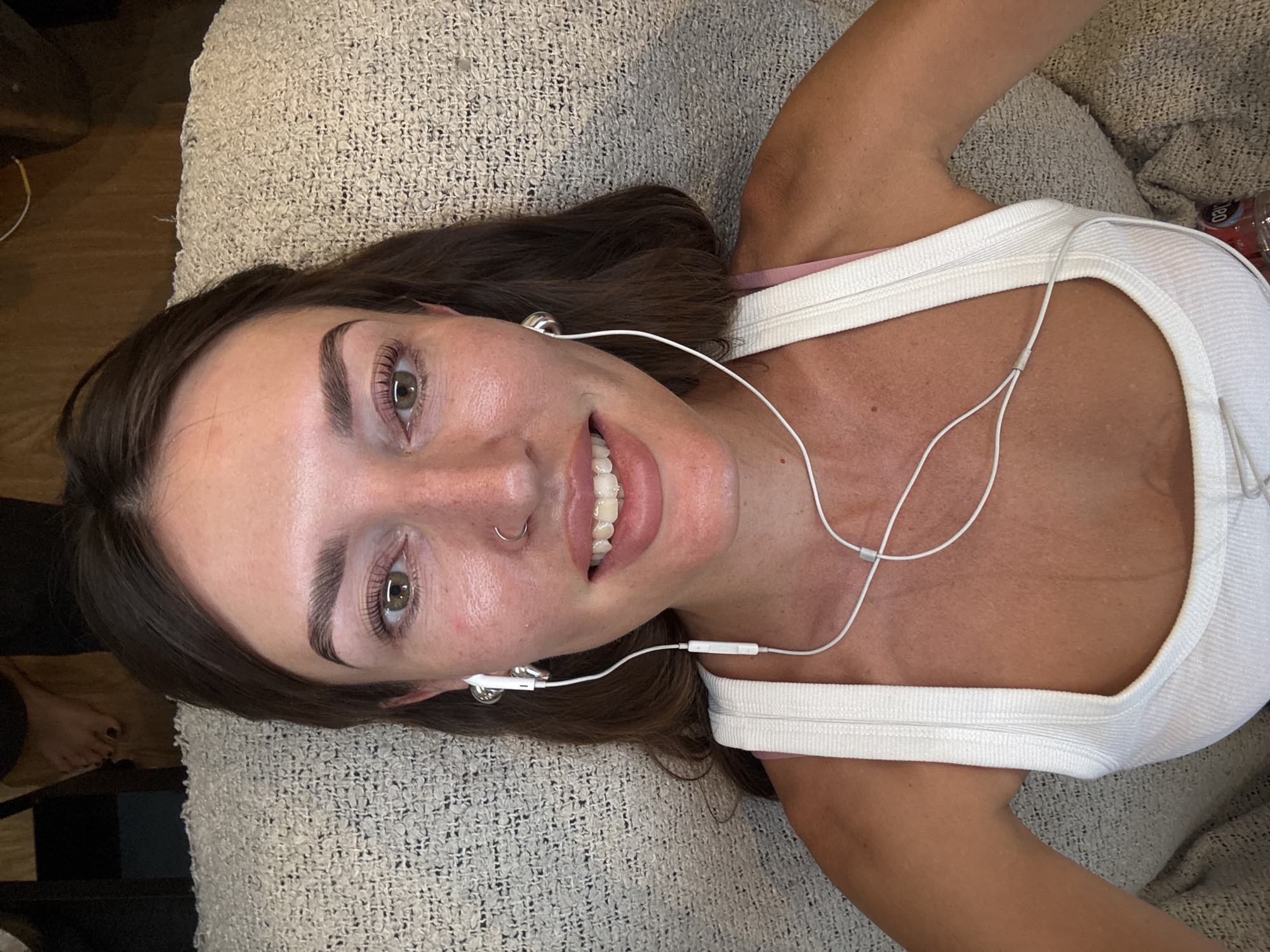 A medium shot captures a woman laying on what appears to be a beige, textured couch or cushion. Her head is positioned towards the top left, and her body extends diagonally across the frame towards the bottom right. She is wearing a white tank top with a narrow strap and appears to be wearing earphones. Her hair is dark brown, and her skin tone suggests fair complexion. She has a nose ring on her left nostril. Her eyes are open, and she is smiling slightly, looking towards the camera. A foot is visible in the bottom left corner. There is a bottle of what appears to be water, with a visible label, near the upper right of the frame. The image does not provide enough information to confirm its location as North Kuta, Indonesia.
