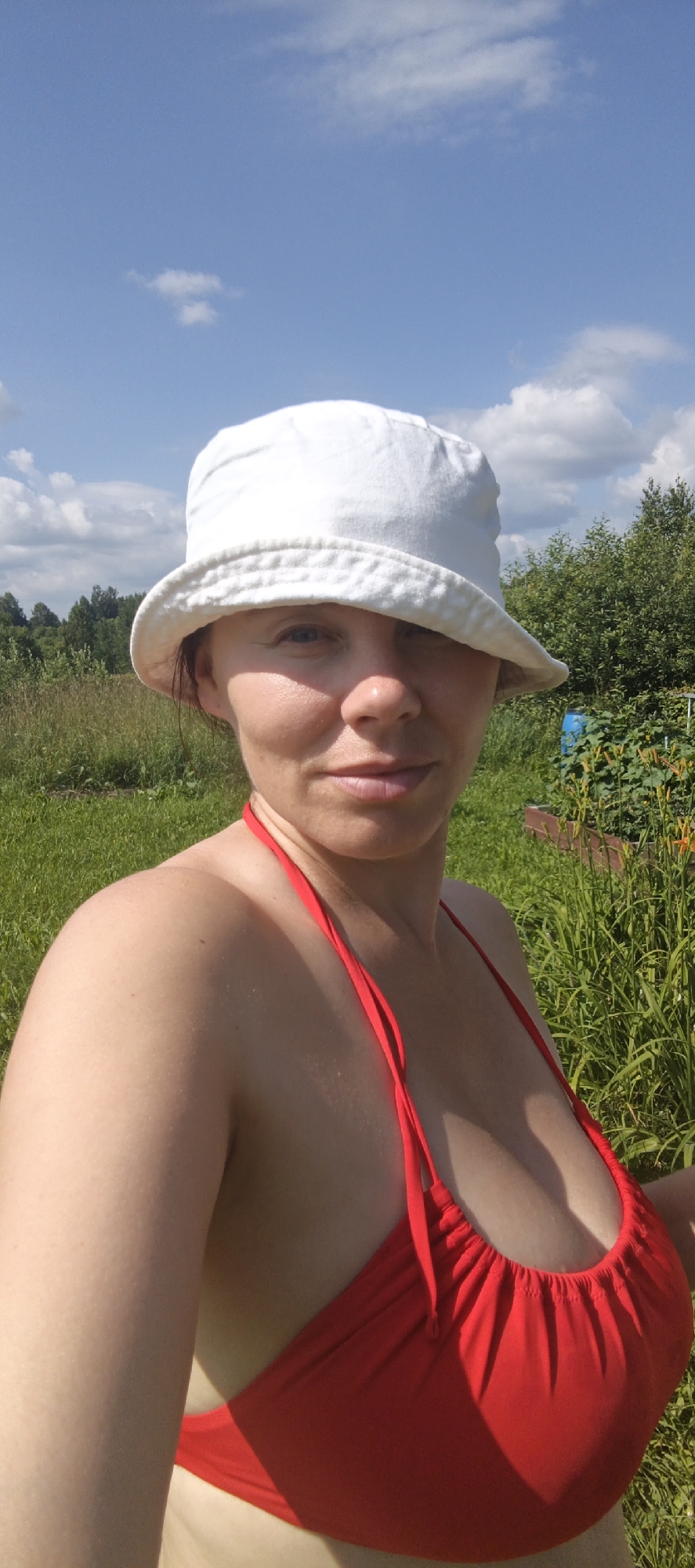 A woman wearing a white bucket hat and a red bikini top is pictured outdoors on a sunny day. She is looking towards the camera. Her skin is fair and slightly tanned. She has light brown hair visible beneath the hat. The background consists of green grass, lush foliage, trees, and a bright blue sky with scattered white clouds. The scene appears to be in a rural or natural setting, possibly a garden or a field, near Большой Букор, Russia. There is a blue barrel and some plants visible in the background. The lighting suggests it is daytime.