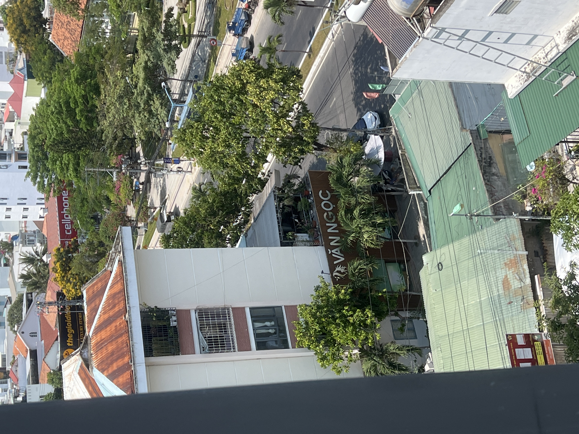 The image captures an elevated view of an urban landscape, likely in Nha Trang, Vietnam. The scene features a mix of residential and commercial buildings of varying heights and architectural styles. Some buildings have pitched roofs covered in red tiles, while others are flat-roofed with what appear to be corrugated metal sheeting or concrete. The building in the foreground is painted white with brown trim and has barred windows. A large banner on another building reads "VĂN NGỌC" in bold letters. Streets run through the scene, and some include pedestrian and vehicular traffic. A notable feature is the presence of lush vegetation, including trees and smaller plants. The general environment seems sunny and bright, suggesting a daytime shot.
