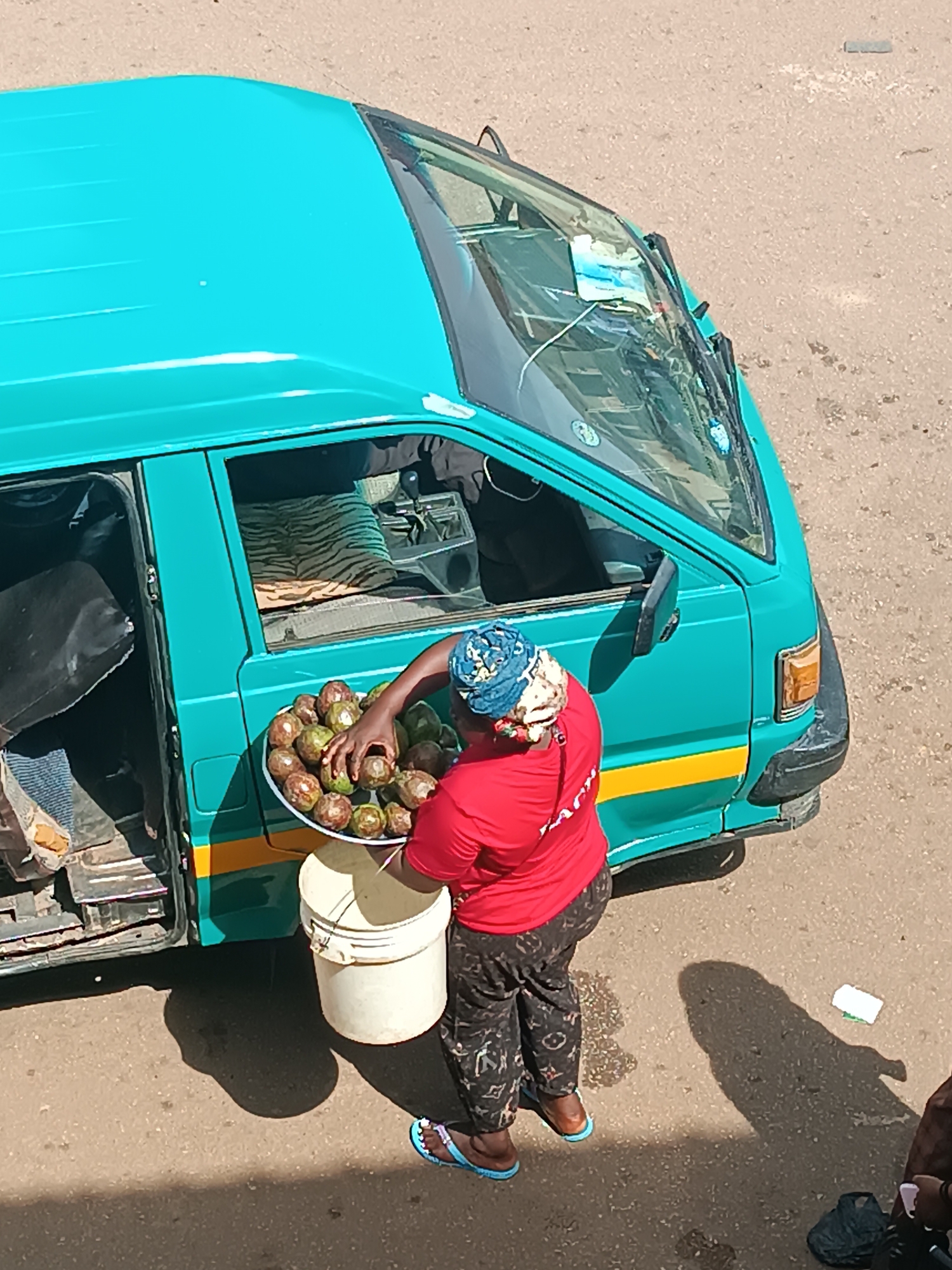 The image depicts a street scene in Bukuru, Nigeria, featuring a woman standing next to a turquoise minivan with an open side door. The woman is wearing a red shirt, patterned pants, and a blue patterned head wrap. She is holding a white platter filled with numerous brown avocados, which she appears to be sorting or selecting from. Beneath the platter, she holds a white bucket. The minivan has a yellow stripe along its side, and the interior is visible, showing patterned seat covers. The ground is paved, and shadows indicate bright sunlight. A discarded white item rests on the pavement. Additional indistinct people appear at the edge of the frame.