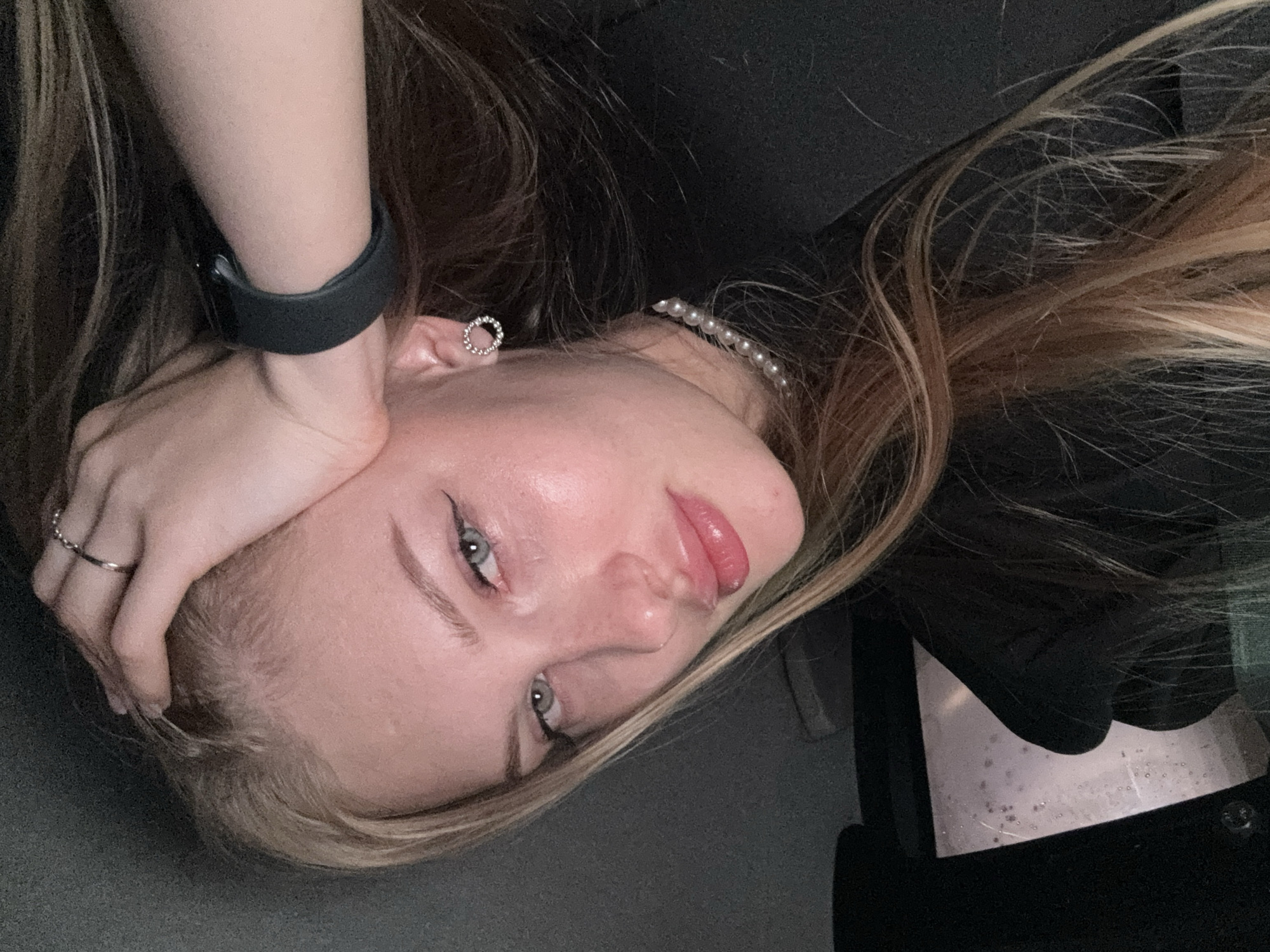 The image shows a close-up of a young woman with blonde hair, blue eyes, and fair skin. She is wearing a black top, a pearl necklace, a ring on her right hand, and a black smartwatch on her left wrist. Her hand is resting on her head. She has on winged eyeliner and lip gloss. The background is dark and out of focus. There is a window with water droplets on it to the left of the woman. The setting appears to be indoors, possibly in a car, as suggested by the window. There is no visible indication of the city of Perm, Russia.
