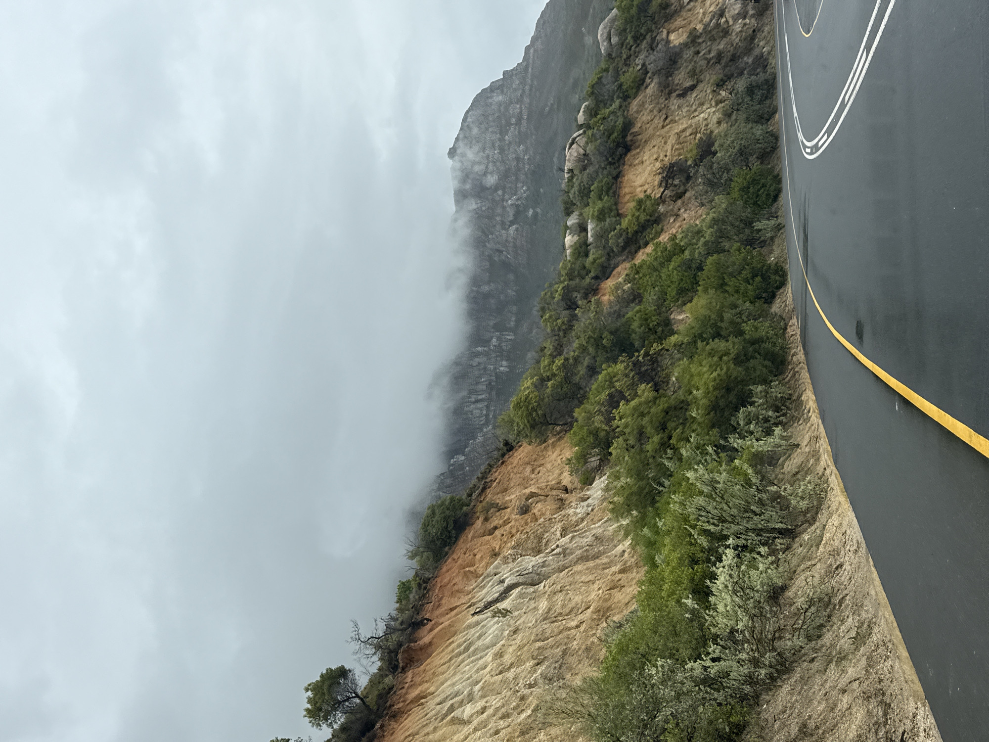 The image captures a scene along a road in Cape Town, South Africa, possibly near Table Mountain. The landscape is characterized by a mountainside with a combination of rock formations, vegetation, and exposed geological layers. A paved road runs alongside the mountain. Overcast skies with low-hanging clouds create a muted and somewhat hazy atmosphere. The road surface is wet, reflecting the light. The road has painted white lines and a yellow line marking its edge.
