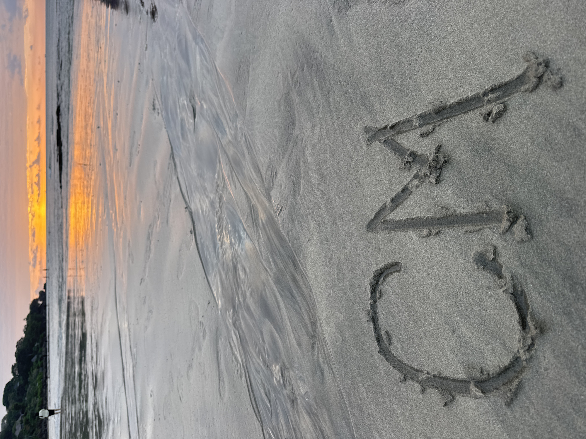 The image depicts a beach scene at sunset in Kuta Selatan, Indonesia. The camera is angled downwards, focusing on the wet sand in the foreground where the letters "CV" are drawn. The sand shows patterns of water runoff, indicating that the tide has recently receded. In the background, the ocean meets the horizon under a sky filled with warm sunset colors, predominantly orange and yellow hues, reflecting on the wet sand. A dark silhouette of land with lush vegetation is visible on the left side of the image, suggesting a coastline. A small figure, possibly a person, is visible near the vegetation line, adding a sense of scale to the vastness of the beach. The overall mood is serene and peaceful, characteristic of a sunset by the sea. There is no other visible text in the image besides the drawn letters "CV".