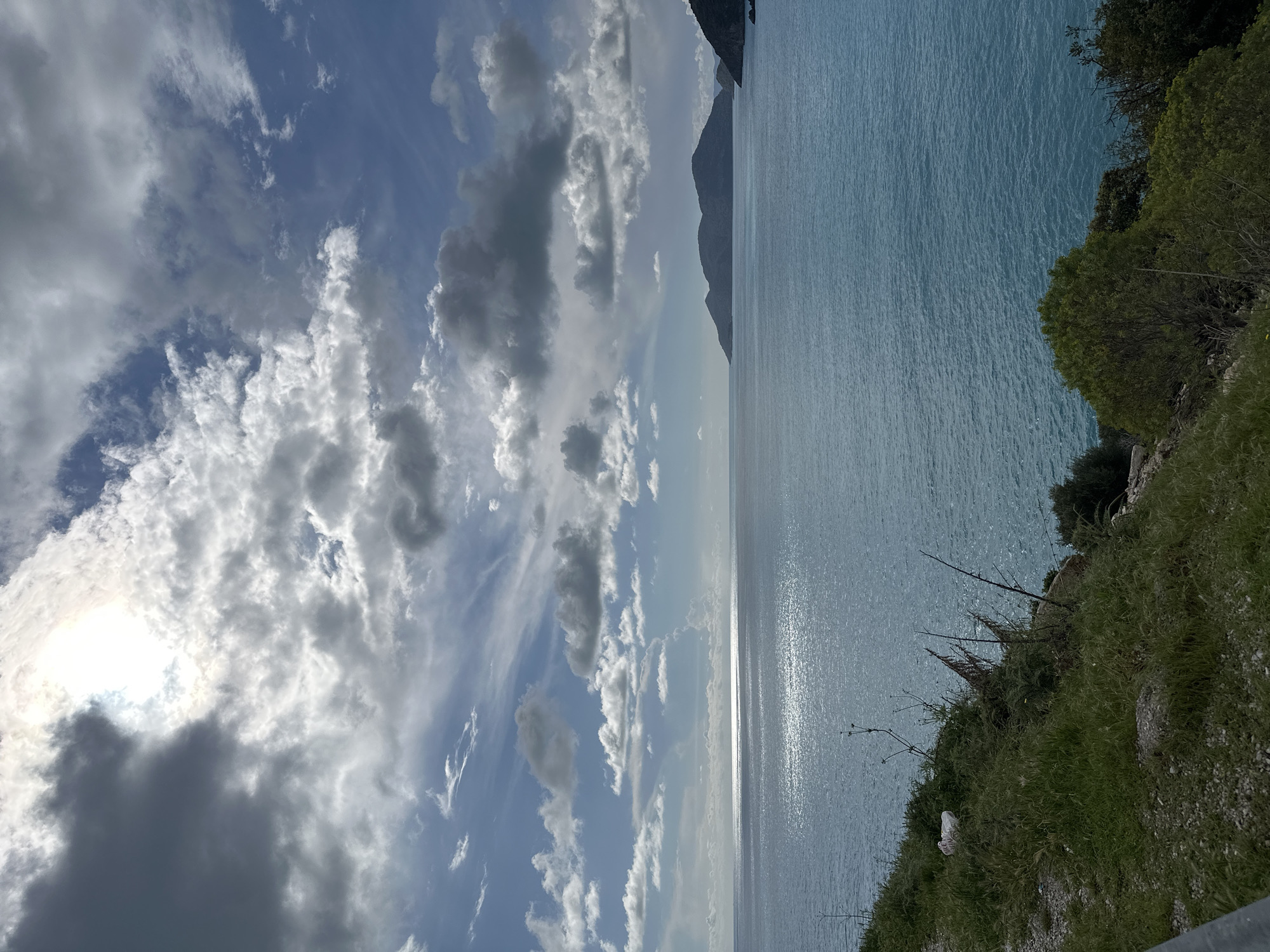 The image shows a coastal scene in Fethiye, Turkey. The sky is filled with partly cloudy skies with a visible sun shining through the clouds, while the horizon line features a mountain range. The sea is visible, shimmering from the sunlight. A rocky coastline with vegetation runs along the bottom of the image.