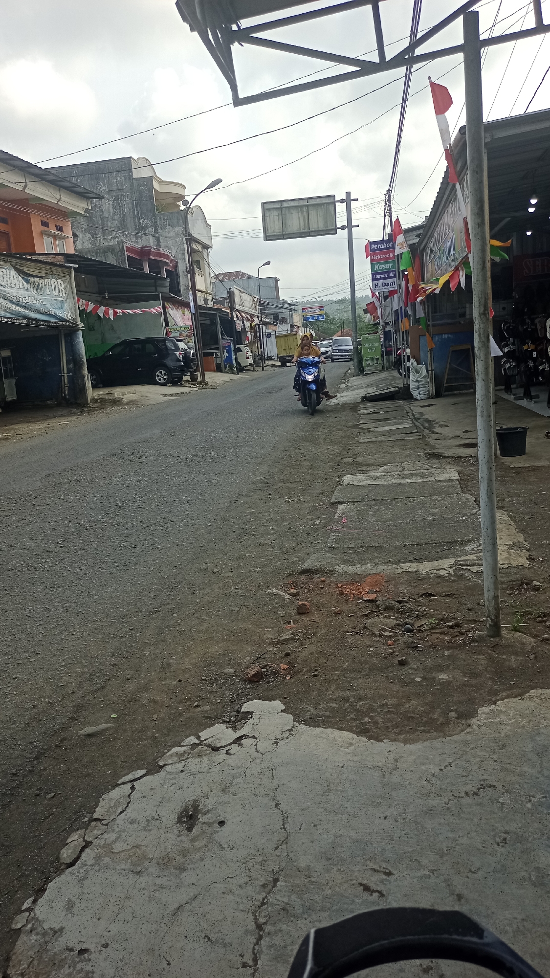 A street scene in Cikalong, Indonesia, features a two-lane asphalt road with a sidewalk on the right. The sky is overcast. On the left side of the road, there are buildings with storefronts, some with awnings. One building has a sign that reads "DIAN MOTOR." A black SUV is parked in front of a building on the left. On the right side of the road, more storefronts are visible, with Indonesian flags and colorful banners decorating the scene. A person is riding a blue motorcycle in the middle of the road, heading away from the camera. Further down the road, other vehicles and buildings are visible. The ground on the right side of the road is a mix of concrete and dirt, with some patches of broken concrete.