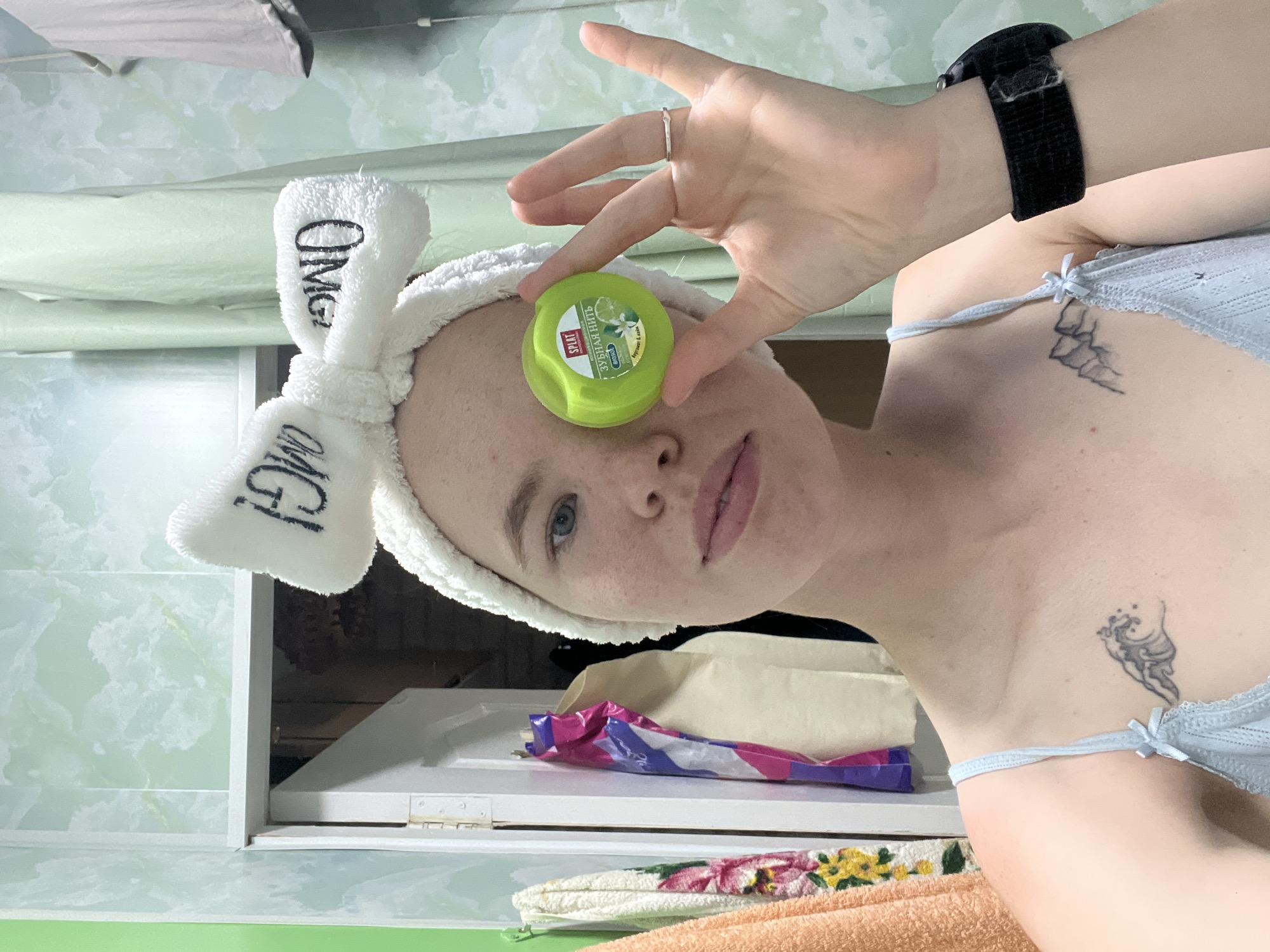 A young woman with light skin, freckles, and blue eyes is holding a bright green spool of dental floss in front of her right eye. She is wearing a white fuzzy headband with black lettering that reads "OMG" on each ear. The woman has light brown hair and is wearing a light blue camisole. A small wave tattoo is visible on her chest. She is indoors in what appears to be a bathroom, indicated by the tiled walls and a towel rack visible in the background. The woman is looking directly at the camera.