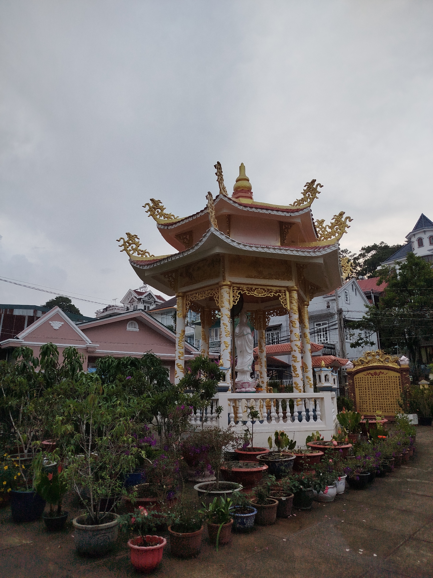 Captured in the city of Xuân Hương, Đà Lạt, Vietnam, the image depicts a pagoda-style structure in a garden setting. The structure is adorned with golden dragons along the eaves and houses a white statue within. Rows of potted plants, varying in size and species, line the ground around the structure. In the background, residential buildings of various colors and architectural styles are visible under a cloudy sky.