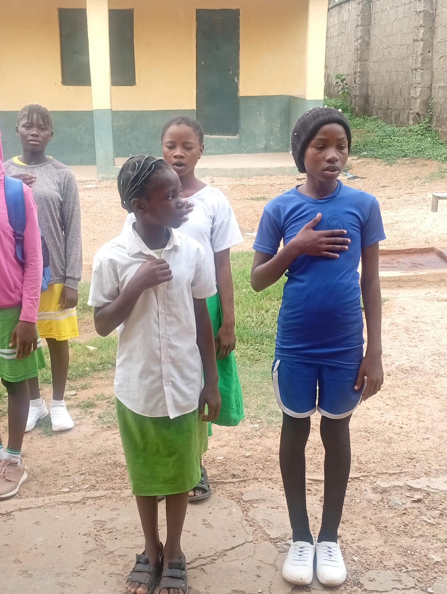 In Narayi, Nigeria, a group of children stand outdoors on a dirt surface, possibly a schoolyard. One child wears a blue t-shirt and shorts with white shoes and a black beanie, standing with their hand over their chest. Another wears a white button-up shirt and a green skirt, also with their hand over their chest, and sandals on their feet. A third child in a white t-shirt and a green skirt also has their hand over their chest. Two other children are partially visible; one wears a grey long-sleeved shirt and a yellow skirt, and the other wears a pink hoodie with a green skirt and a blue backpack. All appear to be reciting something. Behind the children is a building with a beige and green painted exterior, featuring dark green doors and windows. A brick wall and some vegetation are visible in the background.