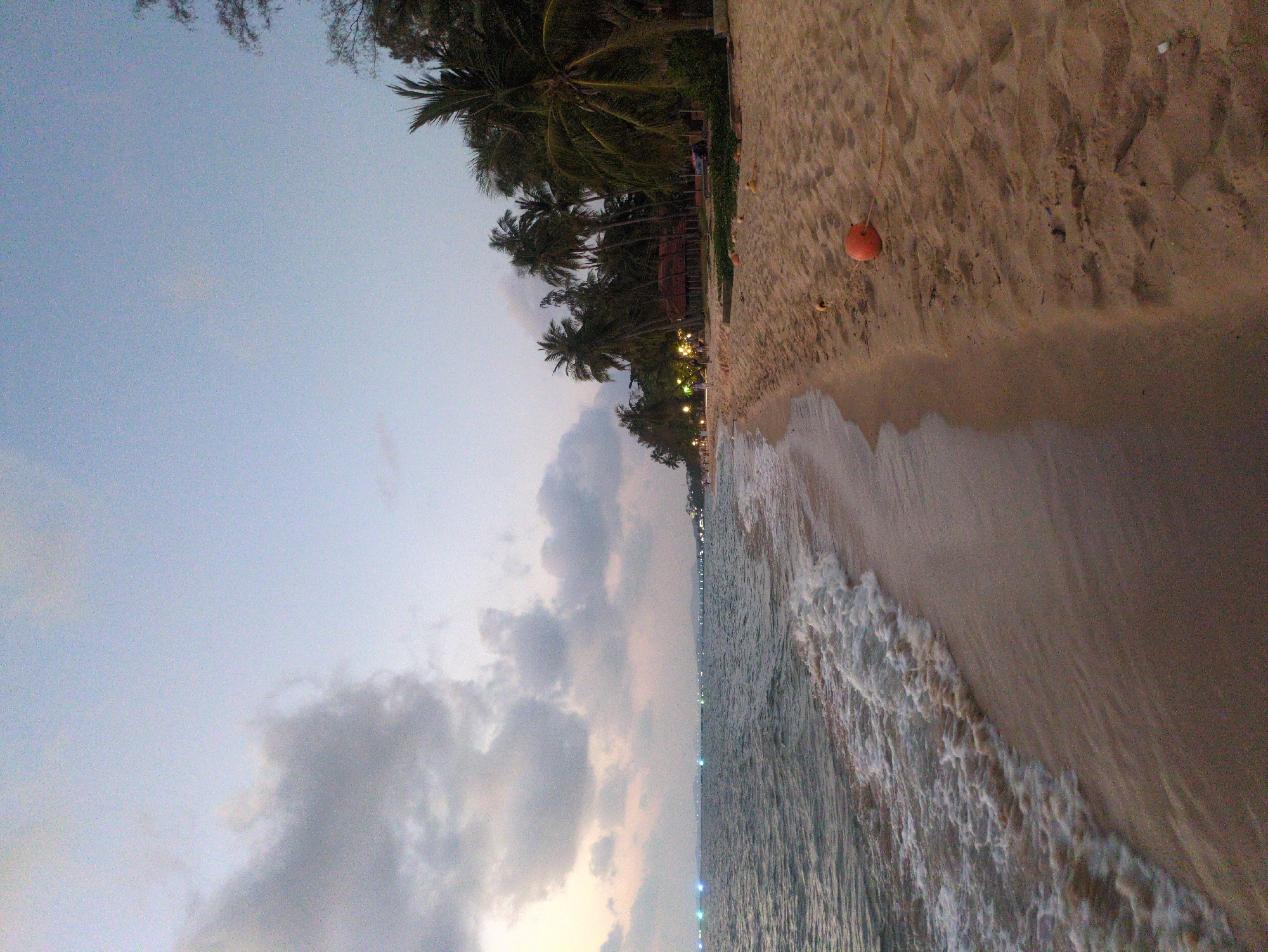 A beach scene, potentially in Phú Quốc, Vietnam, shows a shoreline with palm trees, sand, and waves. An orange buoy sits on the sand. The sky is overcast with clouds. Buildings are visible in the distance.