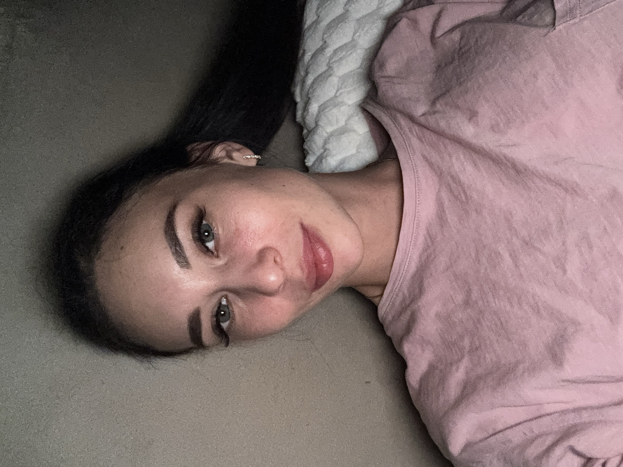 A woman with dark hair is lying down. She is wearing a pink shirt and resting her head on what appears to be a grey surface next to a white quilted pillow. She is looking at the camera. The location is unknown; Chaikovsky, Russia cannot be confirmed.
