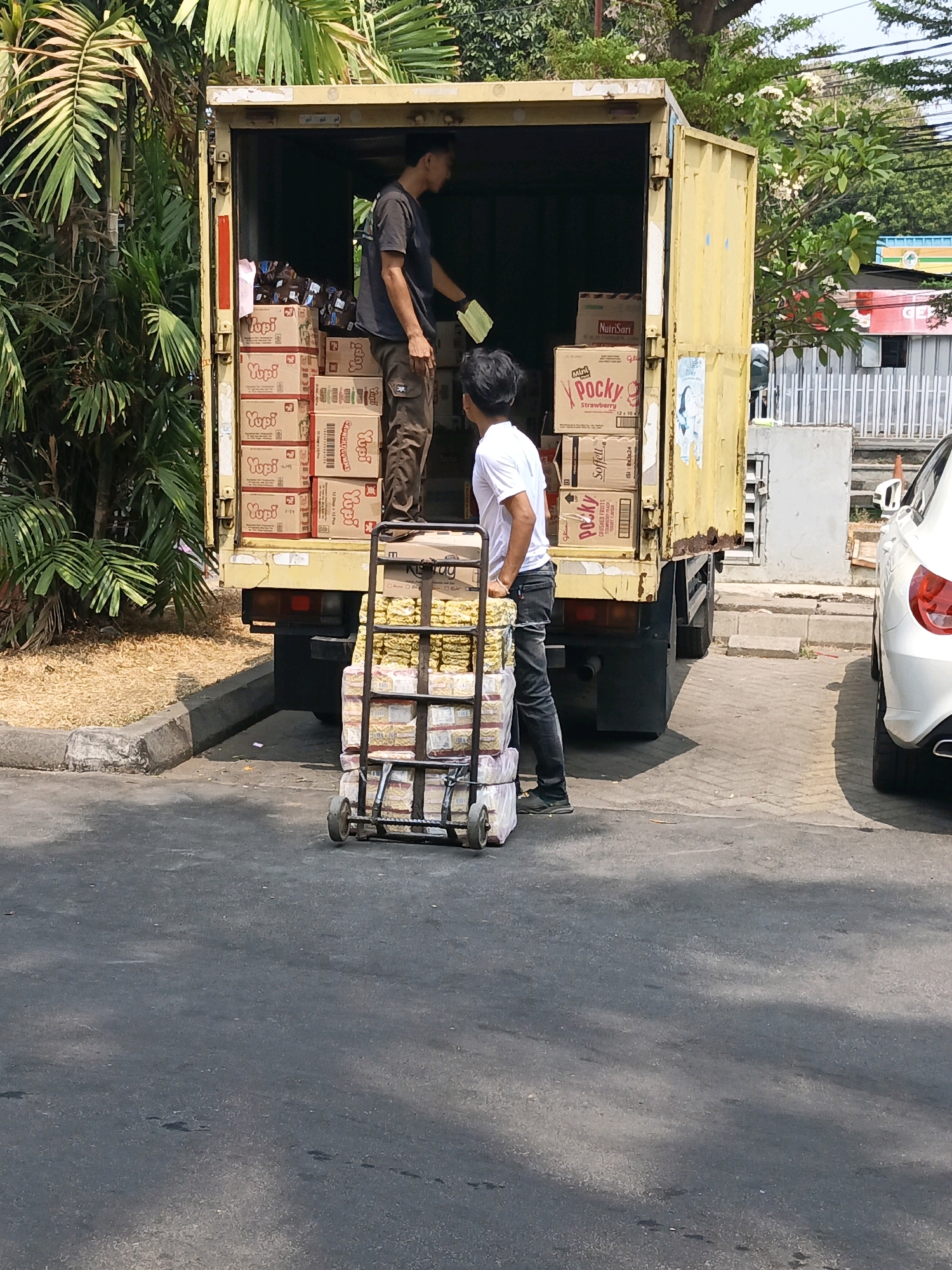 In South Jakarta, Indonesia, two men are unloading goods from the back of a yellow truck. The truck is filled with stacks of boxes, some labeled "Yupi" and "Pocky." One man, wearing a dark t-shirt and brown pants, is standing inside the truck and appears to be handing items to another man. The second man, in a white t-shirt and dark jeans, is standing outside the truck and is preparing to load items onto a handcart. The handcart is already stacked with several packages of what appear to be food items. The truck is parked on an asphalt surface, with a curb and some foliage visible on the left side of the image. A white car is parked to the right of the truck. The scene suggests a delivery or logistics operation in an urban setting.