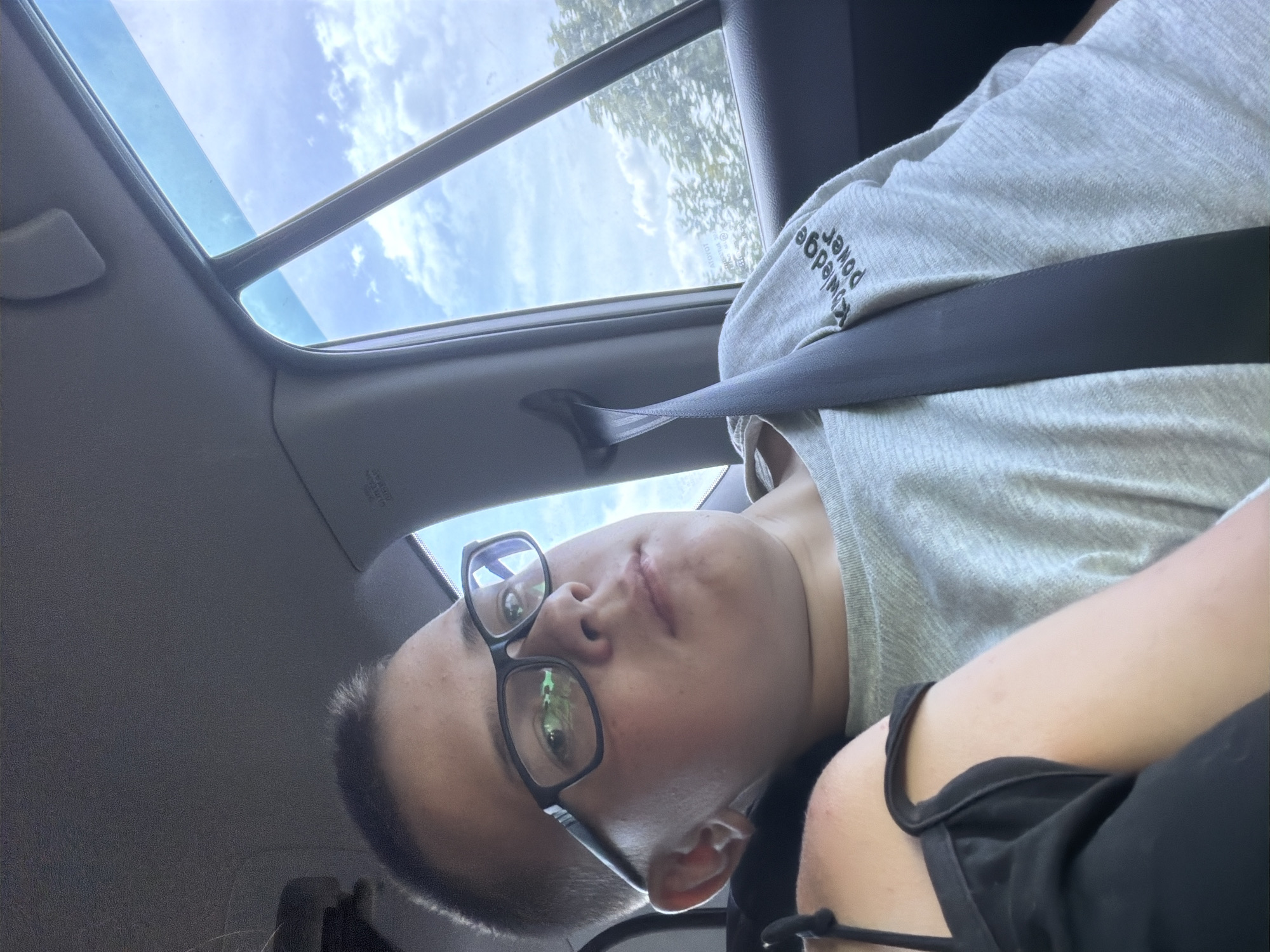 The image shows a young person wearing glasses and a grey t-shirt with "Knowledge Power" printed on it, sitting in the back seat of a car. A seatbelt is visible across their chest. The person is looking towards the camera. The background shows a window with a view of a cloudy sky and trees. The scene appears to be a travel snapshot, but there is no indication of the location being Kruglovka, Russia, or any other specific city.