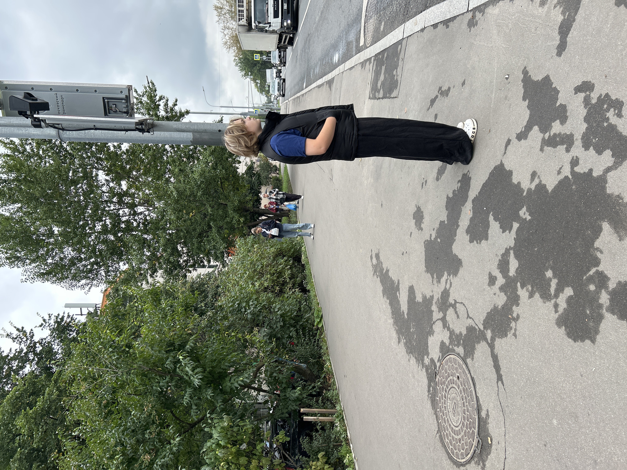 An overcast day in Moscow, Russia, with a young person standing on a sidewalk near a street. The person is wearing a dark vest over a blue t-shirt and loose black pants, along with white Crocs. They are looking towards the street, with their hands in their pockets.
The sidewalk is made of concrete and has some wet patches and shadows. A circular manhole cover is visible on the left side of the sidewalk. To the right of the sidewalk is a road with several cars and a white truck driving on it. A traffic light pole with a camera is also present on the sidewalk.
Behind the sidewalk, there is a row of green trees and bushes, and in the background, parts of buildings are visible. The sky is cloudy, indicating a potentially rainy or damp day. There are other pedestrians in the background, walking along the sidewalk.