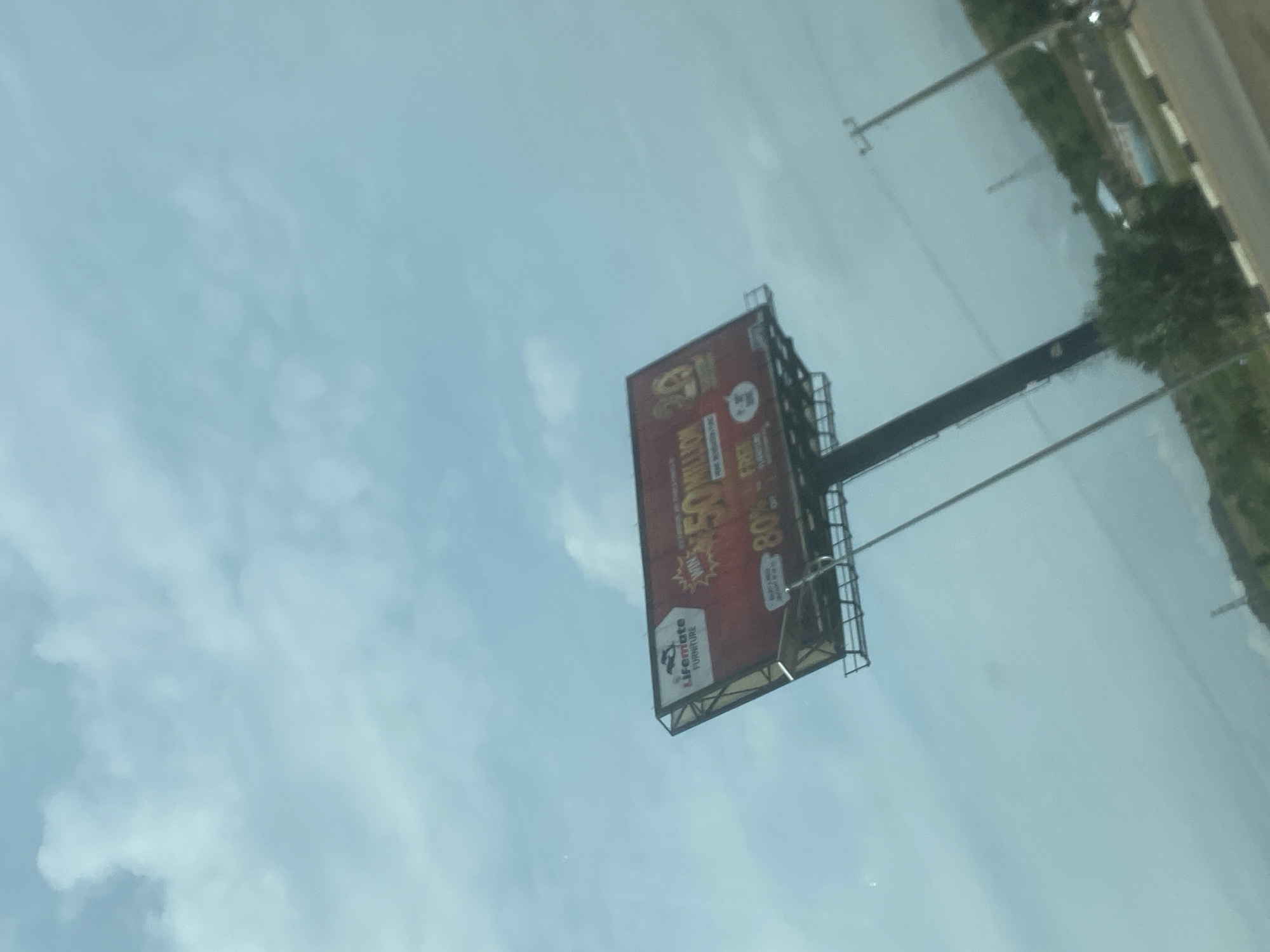 The image captures a large billboard against a bright, partly cloudy sky. The billboard, positioned on the right side of what appears to be a highway or major road, dominates the mid-ground. The sky is a light blue with scattered white clouds, suggesting a clear daytime setting. To the far right, parts of a road, green vegetation, and utility poles with overhead lines are visible, indicating an outdoor, possibly semi-urban or roadside environment.
The billboard itself is rectangular and features a red background with prominent white and yellow text. A logo for "LIFEMATE FURNITURE" is clearly visible on the bottom left of the billboard, accompanied by a graphic of a person seated on a sofa. The main advertisement highlights a "WIN MAXIMUM 50 MILLION" offer, displayed in large, attention-grabbing yellow letters. Below this, another significant offer is advertised as "80% OFF" with additional text below it, including the word "FREE" and some smaller, less legible details. A small circular icon with the number "24" is also present on the billboard.
The scene depicts commercial advertising along a thoroughfare under favorable daylight conditions, with no specific human activity or people visible. The image is tilted, suggesting it was captured from a moving vehicle.