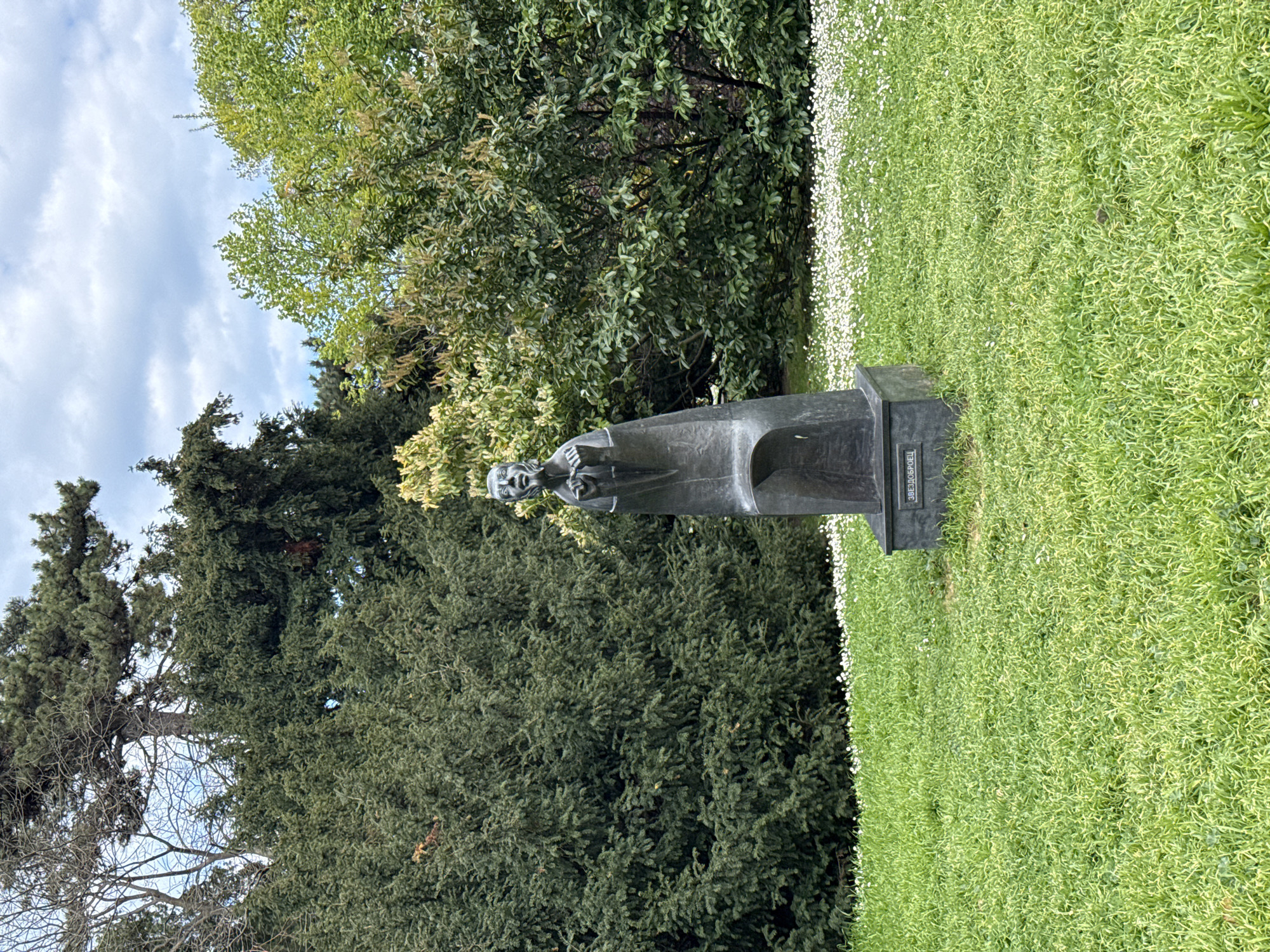 The image shows a bronze statue in a park setting. The statue depicts a male figure in a flowing robe, with a beard and expressive face. It stands on a rectangular pedestal with Cyrillic inscription. The surrounding area is lush with green grass and white flowers, suggesting a well-maintained garden or park. Various trees and foliage are in the background, adding depth to the scene. The sky is partially visible with scattered clouds. The statue is located in the Sea Garden of Varna, Bulgaria.