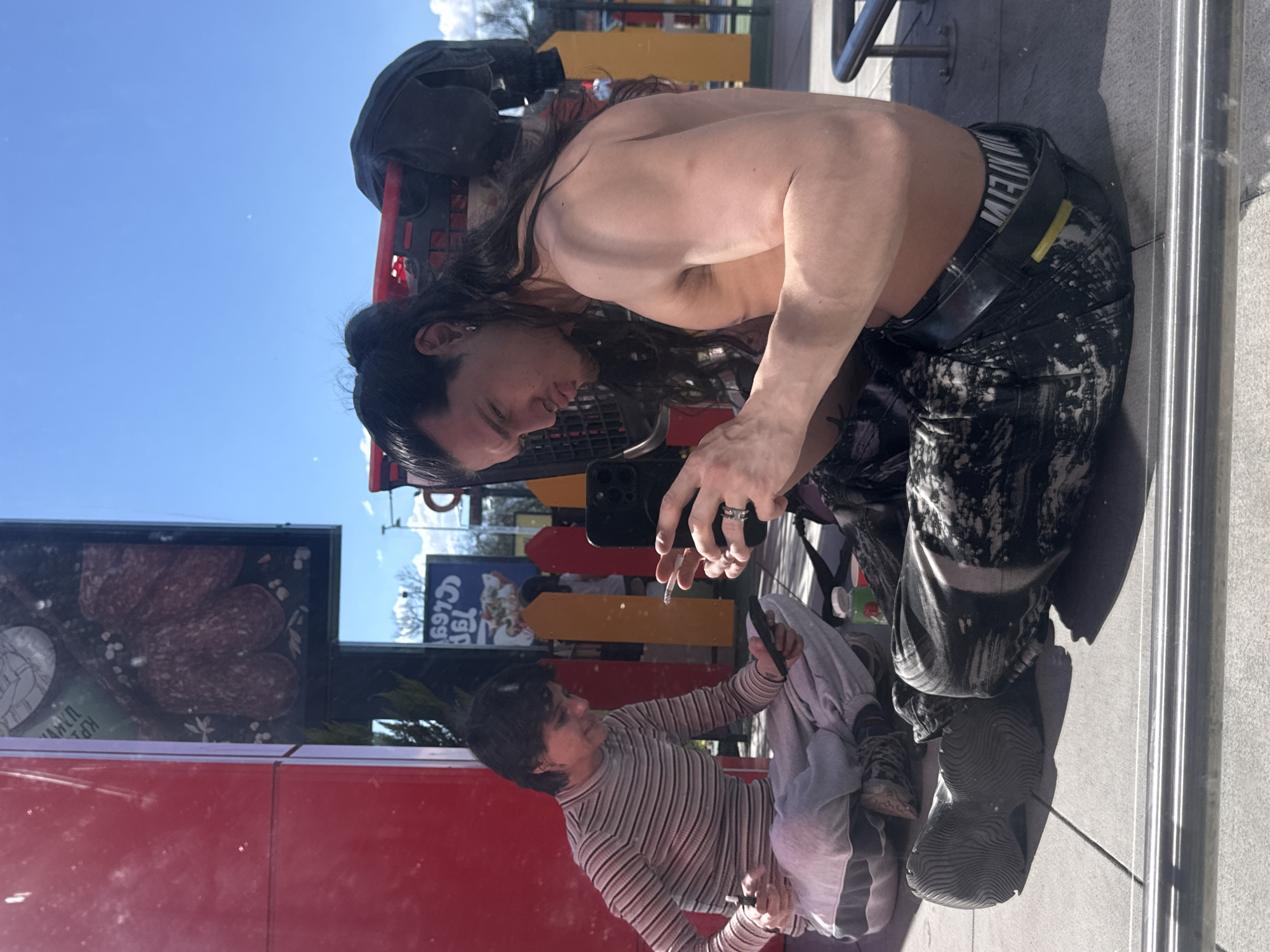 A shirtless person with long dark hair is photographed in Sofia, Bulgaria. The individual is kneeling, holding a smartphone, and smoking a cigarette. They are wearing dark pants with white splatters and a black belt with white lettering. The reflection in a shop window shows the person wearing a striped shirt. The window also displays a food advertisement and products such as sliced meat. Outside, the sky is bright blue. A shopping cart is visible behind the person. The setting appears to be an urban environment.