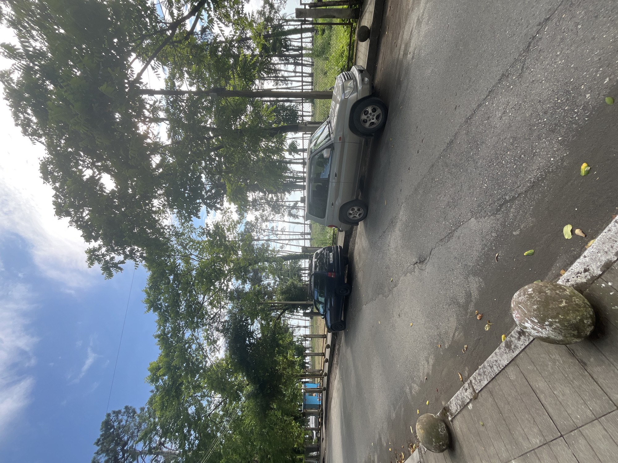 This image depicts an outdoor scene with two cars parked on the side of an asphalt road. A silver SUV is parked in the foreground, and a dark-colored vehicle is parked behind it. Both vehicles are positioned parallel to the curb. The road appears to be wet, and there are some fallen leaves scattered on its surface.
Beyond the road, there is a sidewalk with a wooden plank finish. Two large, spherical, moss-covered stones are placed on the sidewalk near the road, serving as bollards. A line of trees with lush green foliage lines the area behind the sidewalk, obscuring a view of what lies further back. Sunlight filters through the leaves of the trees, creating a dappled effect. In the distance, a fence is visible, and beyond that, more trees and a bright sky with scattered clouds.
The image appears to be taken during the daytime, with bright sunlight indicating fair weather. The presence of trees and the parked cars suggest a roadside location, possibly in a residential or commercial area. The overall atmosphere is calm and serene. There is no visible text in the image.