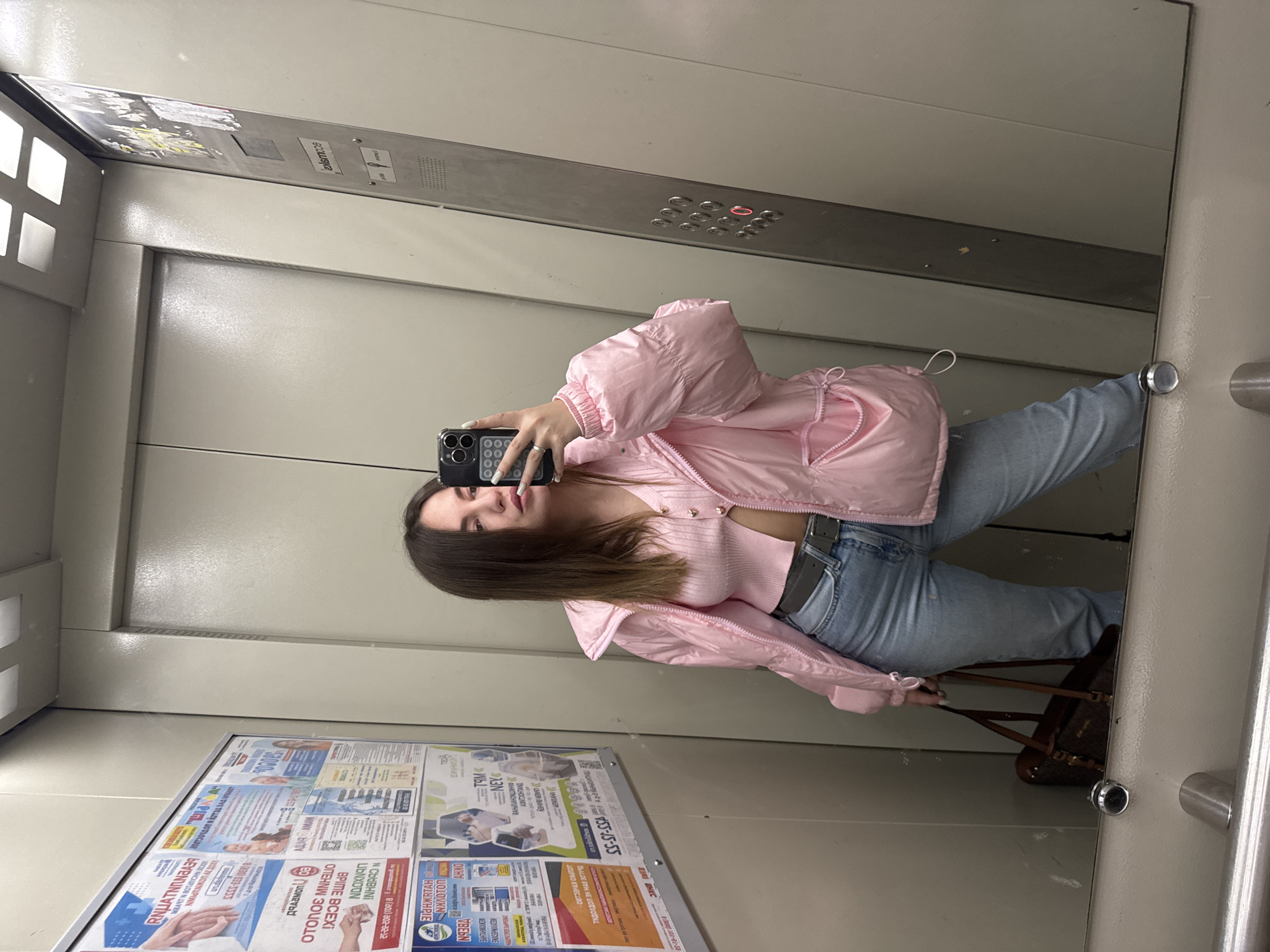 A woman is taking a selfie in an elevator in Orekhovo-Zuyevo, Russia. The elevator has advertisements posted on the wall, including text in Russian and numbers, such as "155-15-55" and "8(800) 101-01-58." The woman is wearing a pink jacket, a matching top, and light blue jeans with a belt. She is holding a phone in her left hand. A brown purse is visible on the floor next to her.