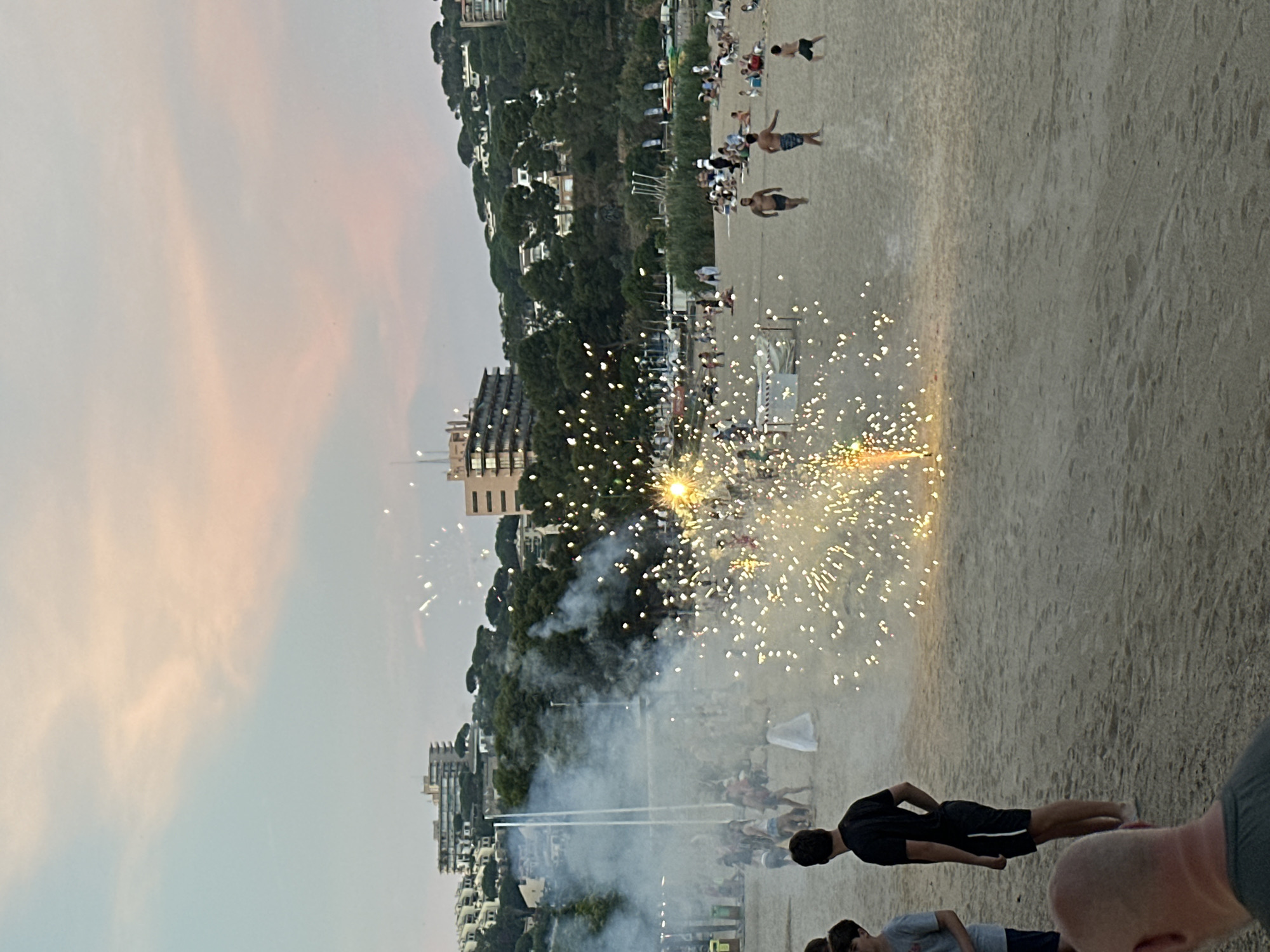 This image captures a beach scene in Sant Feliu de Guíxols, Spain, during what appears to be an evening event featuring fireworks. The beach is populated with numerous individuals, some visible in the foreground and others further back, enjoying the spectacle. Several people are walking on the sand, while others are seated or standing in groups, observing the fireworks display.
In the mid-ground, a prominent fireworks display is in progress, emitting bright sparks and smoke that illuminate the beach. The smoke billows upwards and outwards, creating a hazy atmosphere. To the left of the fireworks, there are tall vertical structures, possibly part of the display or temporary installations for the event.
The background of the image reveals the coastal landscape of Sant Feliu de Guíxols. Buildings, likely hotels or residential complexes, are visible perched on a tree-covered hillside overlooking the beach. The sky above is a soft blend of pastel colors, indicating either dawn or dusk, with clouds adding texture to the scene. The beach itself is sandy, with subtle patterns indicative of foot traffic and activity. The overall impression is one of a lively public gathering on a beach, enhanced by the visual drama of fireworks.
