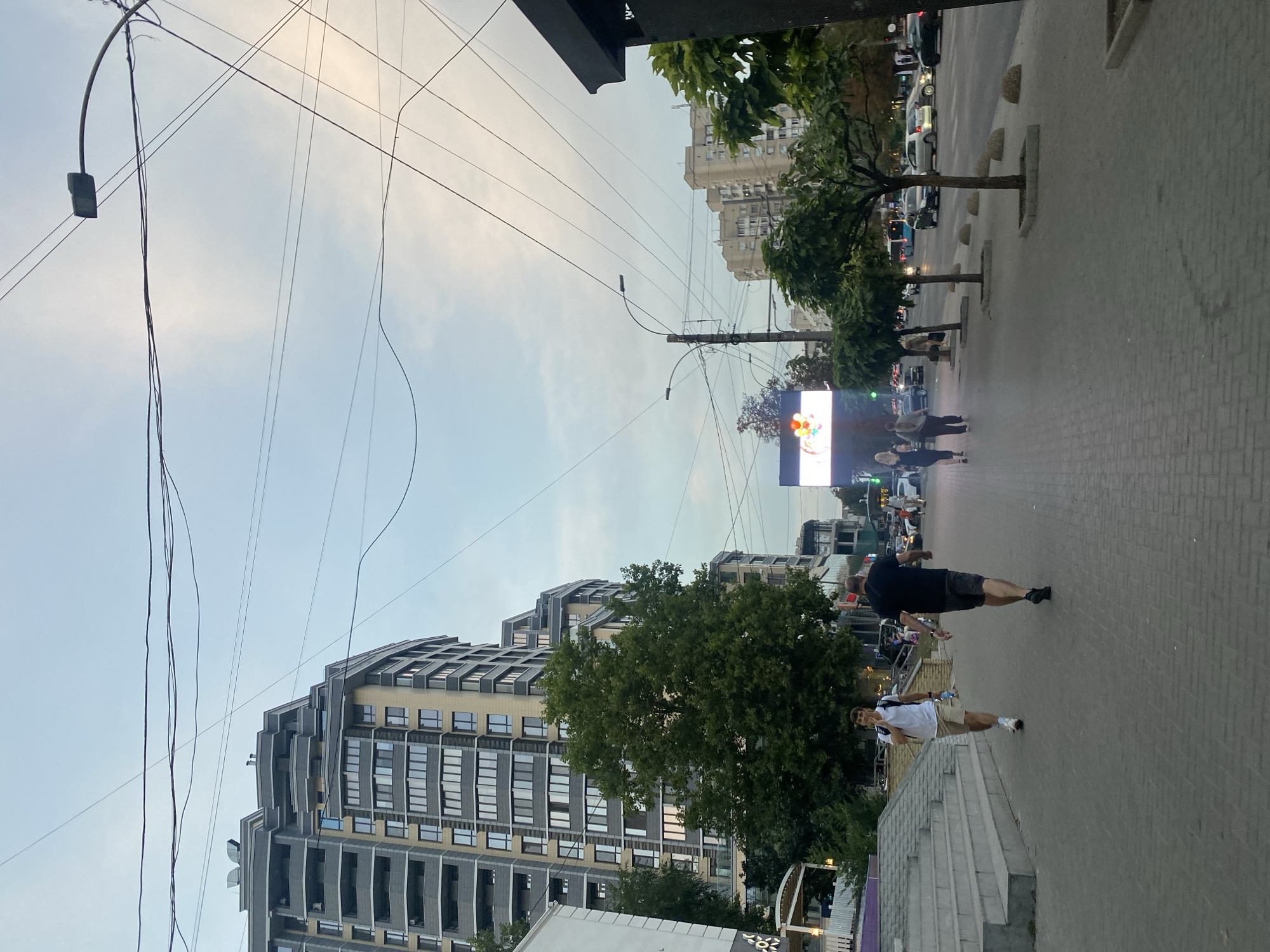 The image depicts a street scene in Chișinău, Moldova, likely during the late afternoon or early evening, given the soft lighting. A wide, paved sidewalk dominates the foreground, with a man in a white t-shirt and khaki shorts walking towards the camera on the left. Behind him, a man in a black t-shirt and dark shorts walks away from the camera. Several other pedestrians are visible further down the sidewalk, moving in both directions.
To the left of the sidewalk, there are concrete stairs leading up to a building. Behind the stairs, a large, modern apartment building with a gray and tan facade stands tall. Lush green trees are interspersed along the sidewalk and street.
Across the street, there is a busy road with several cars and buses. On the right side of the street, more trees line the sidewalk, and another apartment building can be seen in the distance. Overhead, numerous power lines crisscross the sky. A large digital billboard displaying what appears to be colorful balloons is visible on the left side of the street, further down. The overall impression is a typical urban street scene with a mix of modern architecture, greenery, and pedestrian and vehicular activity.