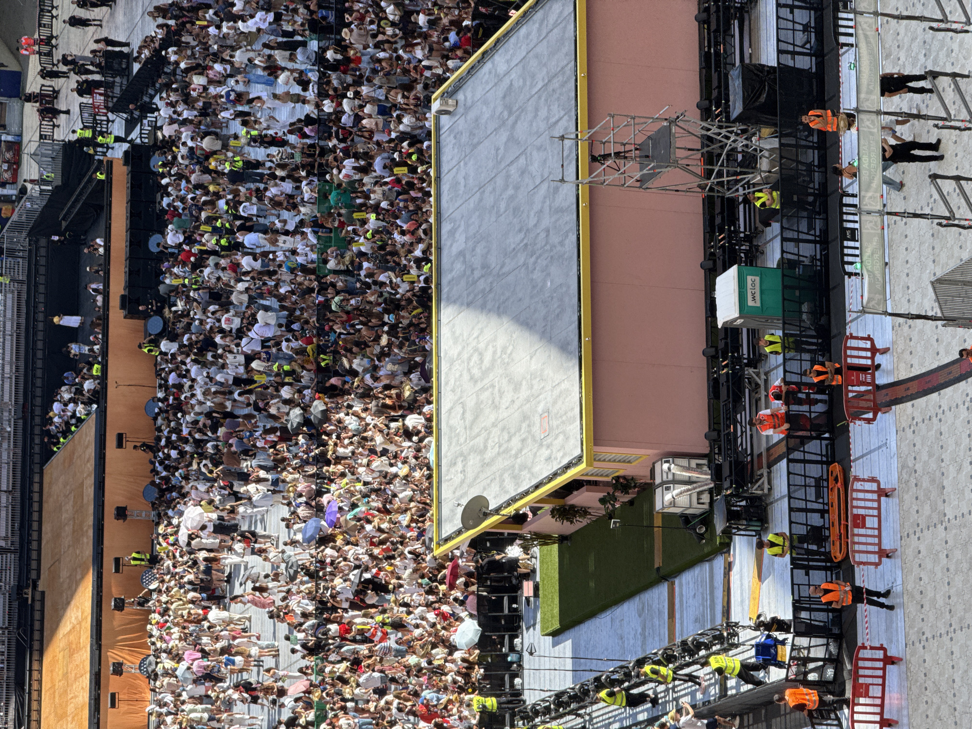 The image presents a high-angle, overhead view of a large outdoor public event under bright daylight. Thousands of people are densely gathered, forming a large crowd that extends across much of the frame. Many individuals in the crowd are wearing light-colored clothing, and numerous attendees are holding open umbrellas, predominantly white or light-colored, suggesting sunny weather and a need for shade. Some people in the crowd appear to be holding yellow signs with visible text like "OFFICIAL."
To the right of the crowd, a significant portion of event infrastructure is visible. This includes a large grey screen with a prominent yellow frame. Below the screen and to its left are multiple stacks of speakers, suggesting a stage or performance area. Numerous event staff or crew members are present, distinguishable by their high-visibility yellow or orange vests. They are positioned around scaffolding, staging, and various equipment. Black tarpaulins cover some equipment, and intricate metal scaffolding structures support lights and other apparatus. Red and orange safety barriers are placed along pathways and structured zones, helping to manage movement.
Specific objects include a green and white portable toilet, clearly labeled "WC LOC," situated within the event setup. A white banner behind some scaffolding reads "SKY," and another banner displays the mirrored text "EMERGENCY." The scene depicts a large-scale gathering, likely a concert, festival, or public viewing event, with comprehensive temporary infrastructure for attendees and performers. The strong sunlight casts distinct shadows, confirming it is daytime.