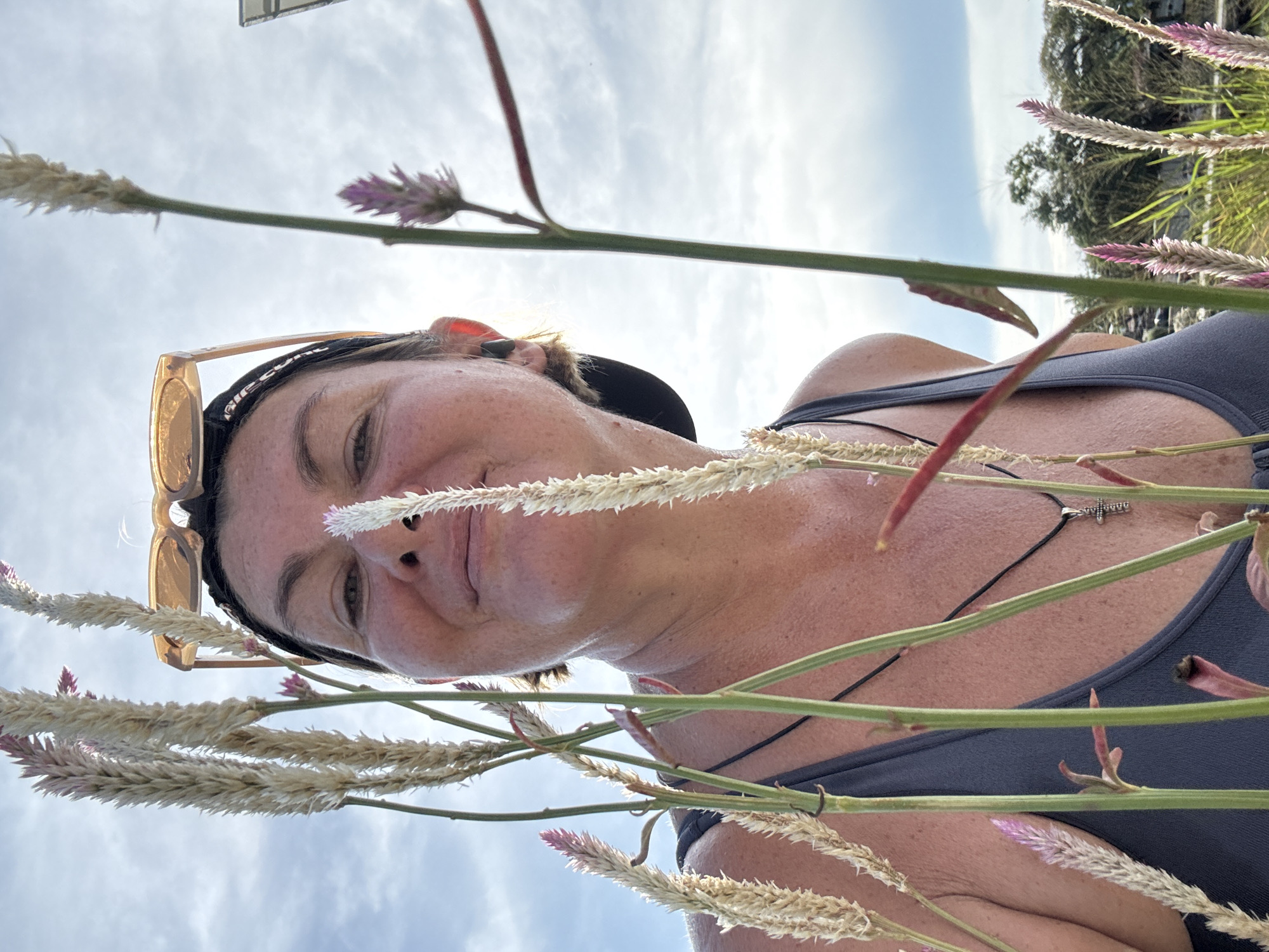 The image depicts a person lying in a field of flowers, looking upwards at the camera. The person has fair skin, a black baseball cap, and sunglasses perched on their head. They are wearing a gray tank top and a black necklace with a cross pendant. The flowers are tall, with feathery, light-colored heads and pink tips. The background shows a blue sky with scattered clouds. The presence of tropical vegetation suggests the location is outdoors and likely in a warmer climate. Given the provided location of บ้านบางรักษ์, Thailand, the scene likely represents a natural area within that region, where the person is relaxing or posing among local flora.