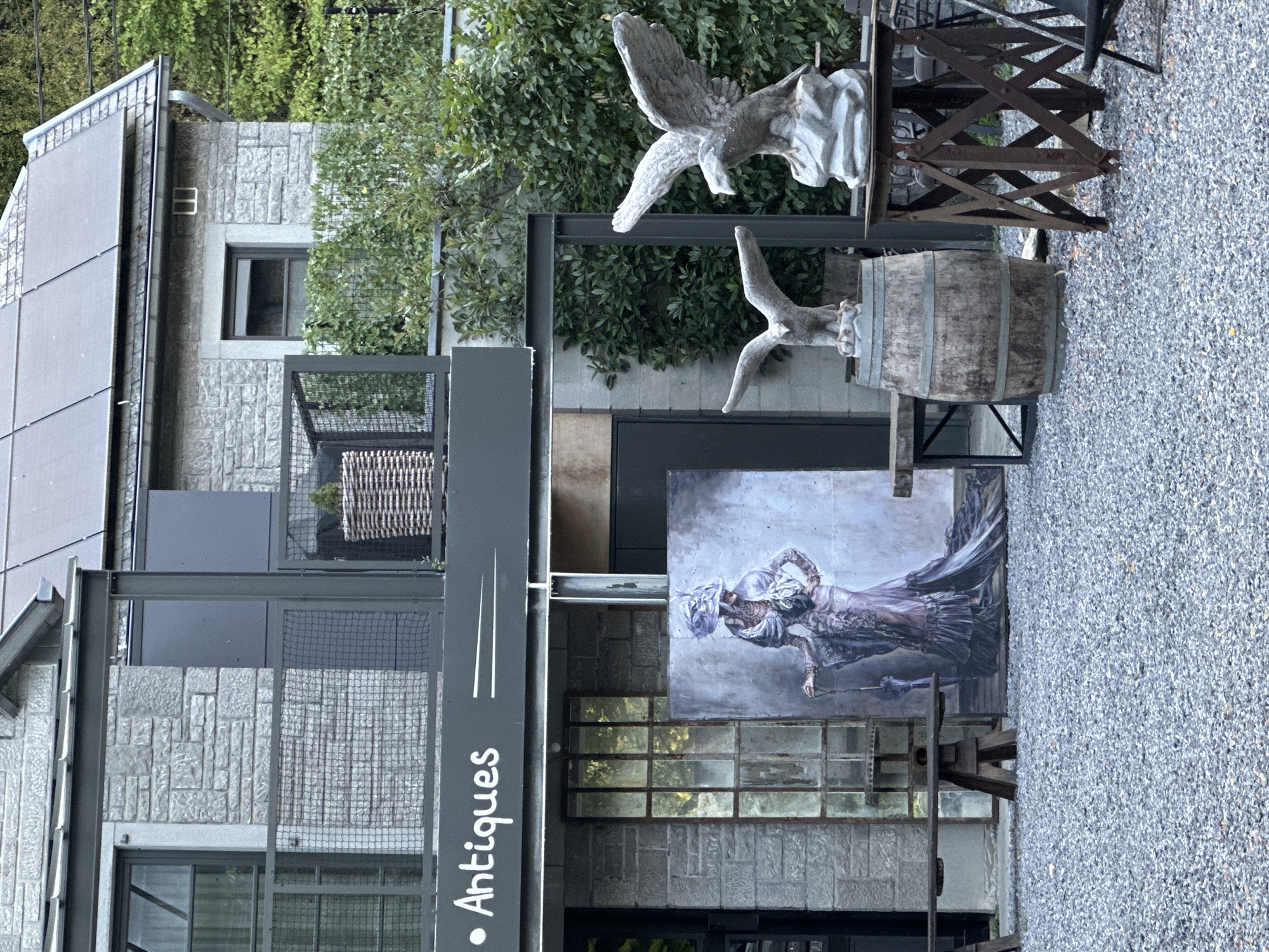 The image depicts the exterior of an antique shop in Durbuy, Belgium. The shop's facade features a stone building with solar panels on the roof. A sign above the entrance reads "Antiques" in white lettering. In front of the shop, there are various items on display, including a large painting of a woman in a vintage dress, a rustic wooden barrel, two eagle sculptures on stands, and a weathered window frame. The ground is covered in gravel. The scene appears to be set in a natural environment with lush green trees visible in the background.