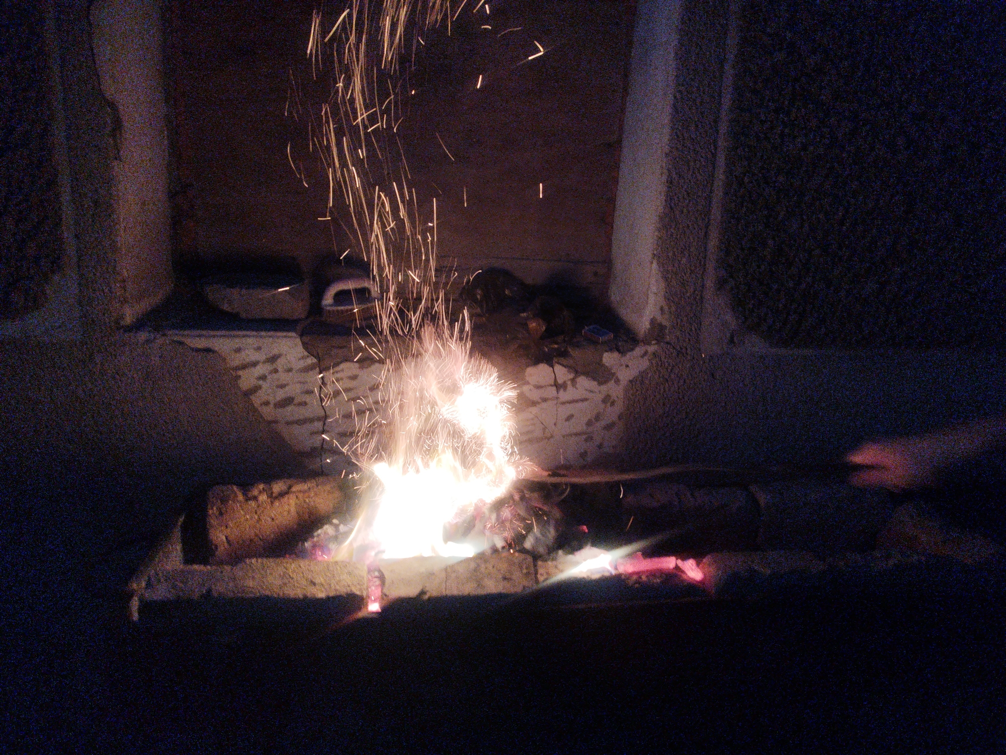 The image depicts a close-up of a fire burning outdoors at night. Sparks are shooting upwards from the flames, which are concentrated on several pieces of wood or charcoal arranged on a rough, possibly stone or concrete surface. The fire is bright and intense, casting a warm glow on its immediate surroundings. Behind the fire, the background is dark and indistinct, suggesting it's nighttime and possibly in an area with limited lighting. There are structural elements visible, which appear to be made of stone or concrete, possibly part of a building or outdoor fireplace. The overall impression is one of a contained, possibly controlled fire. No specific elements indicate the location within Shymkent, Kazakhstan.