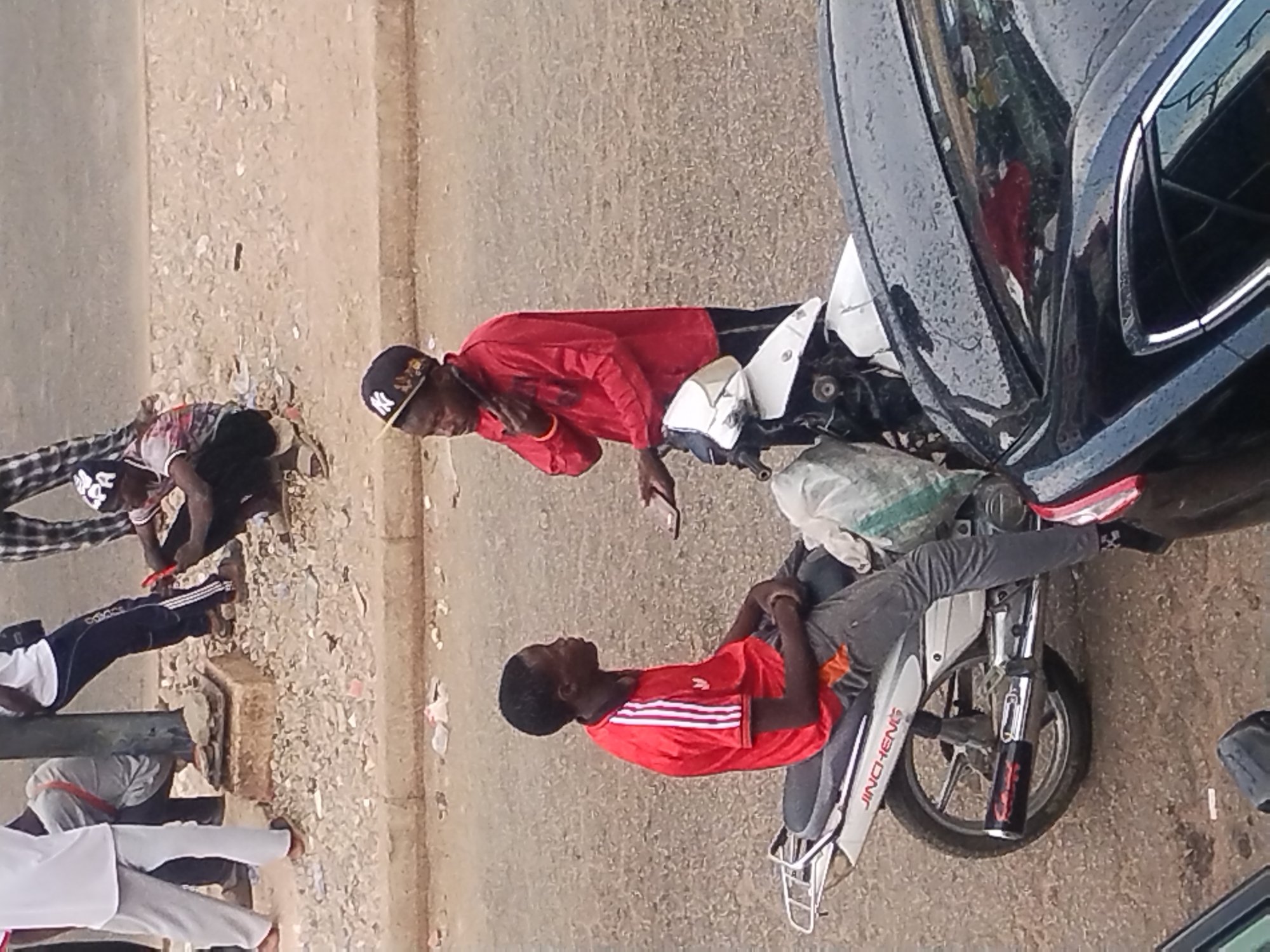The image shows a street scene. Two men in red shirts are sitting on a white motorcycle near a dark blue car. One of the men is wearing a black baseball cap with a logo. Another group of people are near a curb on the side of the street. The location is in a city identified as Unknown, Unknown. The environment appears to be a paved street with some loose debris on the side.