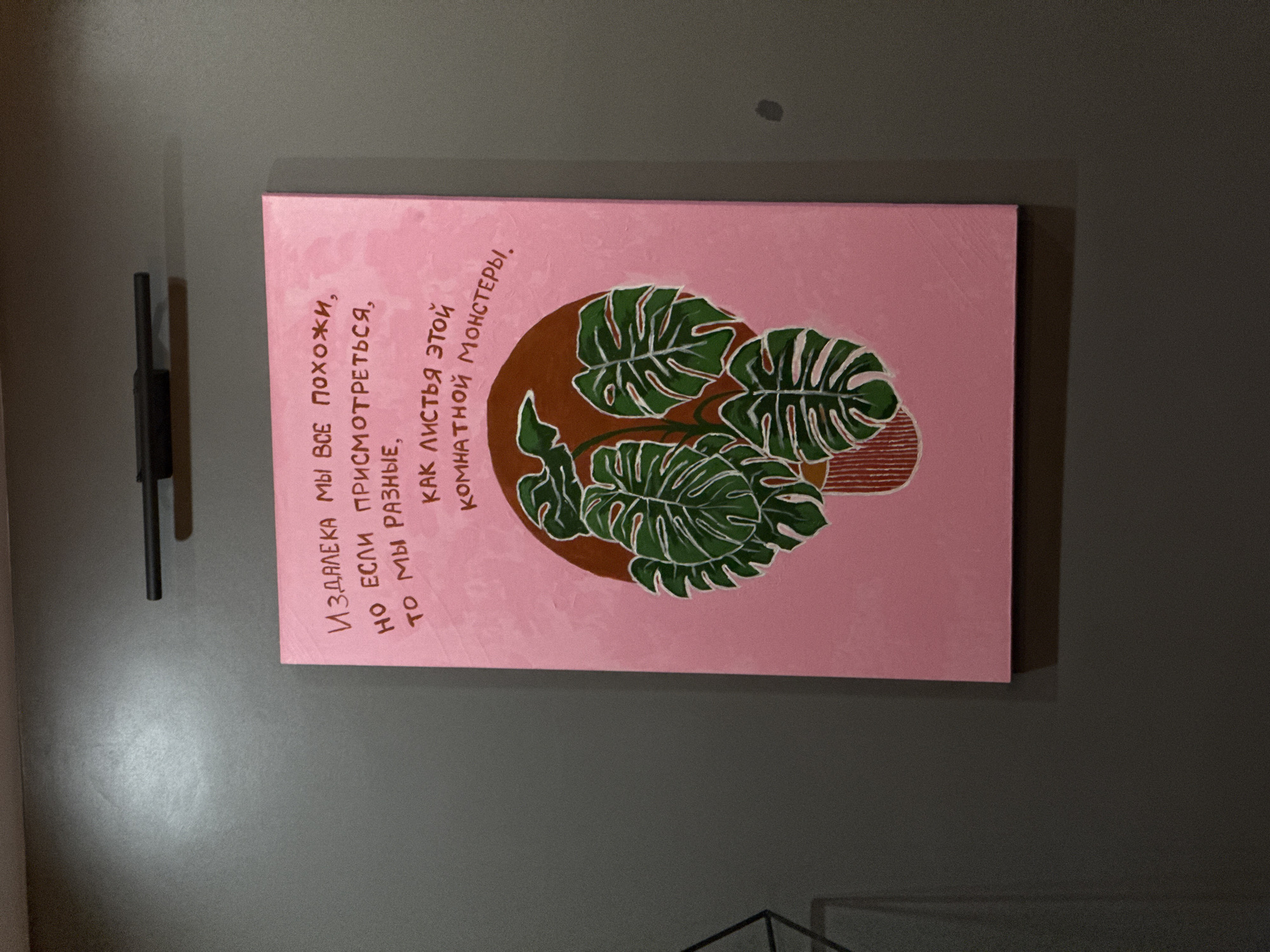 The image shows a painting hanging on a gray wall. The painting is predominantly pink and features a depiction of monstera leaves within a brown circular shape. Text in Russian is present to the left of the monstera leaves. To the left of the painting is a dark, vertical fixture. A small dark spot is visible on the wall above the painting. A corner of a black metal structure is visible in the lower right corner of the image. The image was taken indoors. The location, city, and country are not deducible from the image.