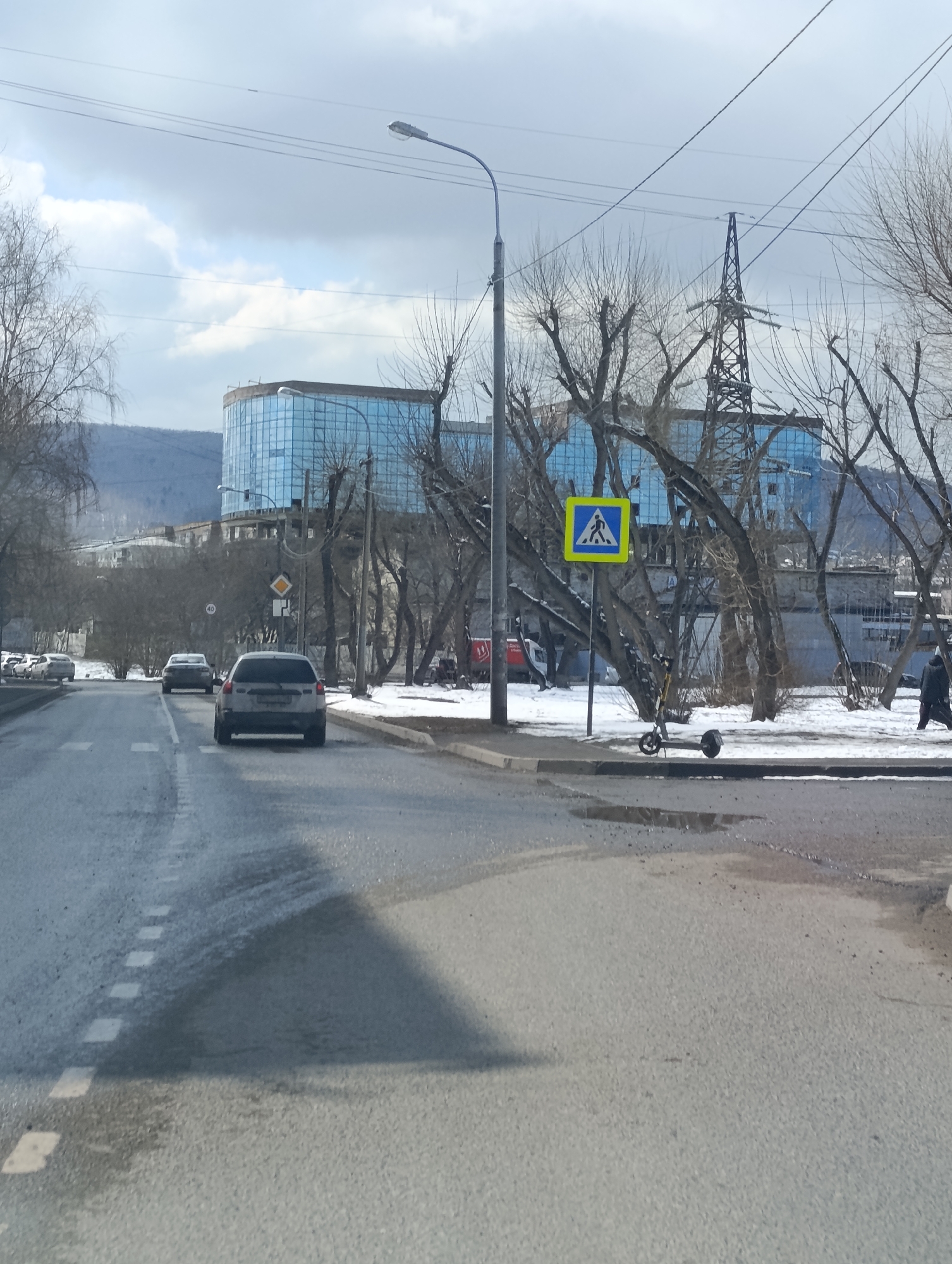 The image depicts a street scene in Krasnoyarsk, Russia, on a cloudy day with bare trees and patches of snow. A wet, asphalt road is visible in the foreground with a car moving away from the camera. A pedestrian crossing sign is located on the right side of the road near a scooter. Across the street, a large blue glass building is visible in the background along with an electrical pylon. Other buildings and trees line the street, with distant mountains in the background. A few people are walking on the sidewalk.