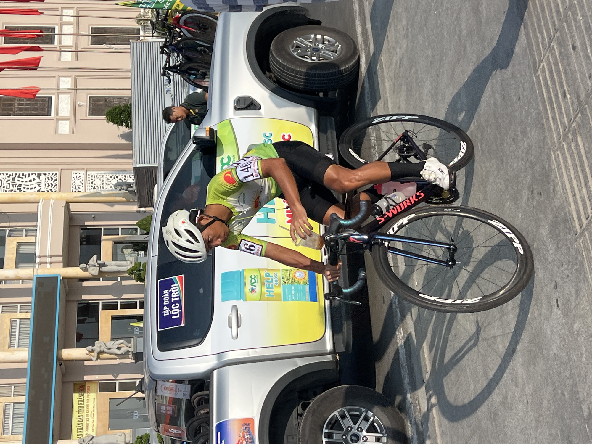 A cyclist is interacting with the passenger door of a white pickup truck during a race in Nha Trang, Vietnam. The cyclist, wearing a helmet, green cycling jersey with the number "146", and black shorts, is bent over, possibly receiving or taking something from a man leaning out of the truck window.
The pickup truck, which has advertisements on its door for "HELP 400SC" and the "Lộc Trời" group, also has bicycles racked in its truck bed. Additional bicycles are stacked vertically on a support behind the truck. The truck is parked on what appears to be a paved road or street. The building in the background is cream-colored with detailed architectural features, including sculptures of human figures. The phrase "AN NHÂN DÂN TỈNH KHÁNH HÒA COMMITTEE OF KHANH HOA PROVINCE 48 Trần Phú, Thành Ph" is partially legible on signage near the building's entrance. The street is bathed in bright sunlight.
