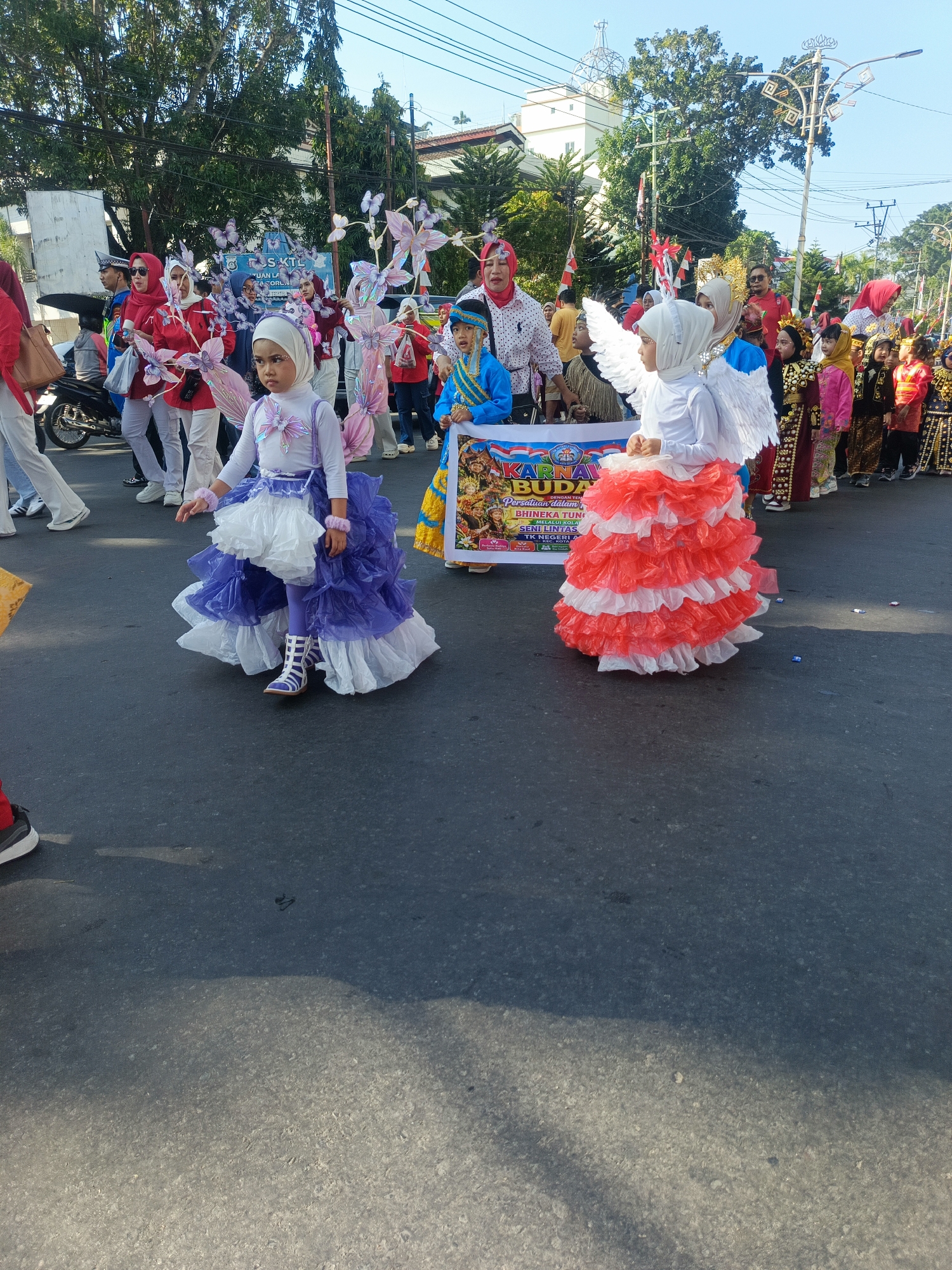 A daytime, eye-level, outdoor, full shot captures a street parade in Gorontalo, Indonesia. The foreground is dominated by two young girls in elaborate costumes. The girl on the left wears a white long-sleeved shirt, a purple and white layered skirt with ruffles, and white and purple striped boots. She is wearing a white hijab. The girl on the right wears a white long-sleeved shirt, a red and white layered ruffled skirt, and a white hijab with attached white wings. She has a colorful banner in front of her that reads "KARNAVAL BUDAYA" which translates to "Culture Carnival." The banner also has text and images related to "BHINNEKA TUNGGAL IKA" (Unity in Diversity) and "SENI LINTAS" (Cross-cultural Art). Behind the girls, other participants in the parade are visible, many wearing colorful traditional Indonesian attire and some holding decorative elements resembling butterflies. The parade is taking place on a paved street lined with trees and buildings under a clear blue sky. A motorcycle is parked on the left side of the image. The overall atmosphere suggests a festive community event.