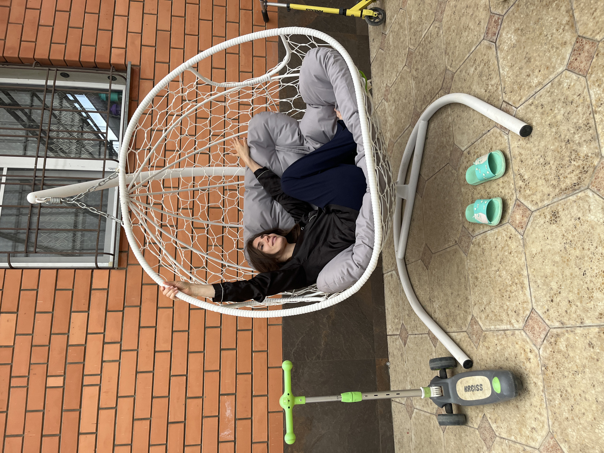The scene depicts a female individual relaxing in a white hanging egg chair within an outdoor or semi-outdoor setting, specifically identified as Kaspiysk, Russia. The woman, appearing to be an adult with dark hair, is positioned on her back, facing right, with her head tilted slightly upward and eyes closed or gazing in a relaxed manner. She is dressed in a dark, possibly black, long-sleeved top and dark blue trousers. Her left arm is extended upwards, grasping the upper rim of the hanging chair, while her right arm rests bent on the light gray cushion.
The chair itself is constructed of white woven material with a distinct egg shape, suspended by a metal chain from a curved white metal stand which rests on the patterned ground. The interior of the chair is fitted with multiple light gray padded cushions providing seating and back support.
The background consists of a reddish-orange brick wall, featuring dark grout lines, spanning the upper half of the image. In the upper right quadrant of this wall, a window with white framing and dark metal security bars is visible. Below the brick wall, a dark gray or brown horizontal band, possibly a foundation or trim, separates the wall from the ground.
The ground surface is paved with geometric tiles, likely hexagonal or octagonal, displaying shades of beige, light brown, and grey, creating a patterned effect.
Several objects are present on the tiled ground:
- To the left of the hanging chair, a grey and bright green three-wheeled scooter with "KIDS" embossed on its deck stands upright.
- To the right, near the base of the hanging chair's stand, a pair of light blue/mint green slides with patterned straps are neatly placed.
- Further to the right, in the background, a yellow two-wheeled kick scooter with black wheels and handlebars is visible.
The scene is illuminated by natural light, indicating daytime.
