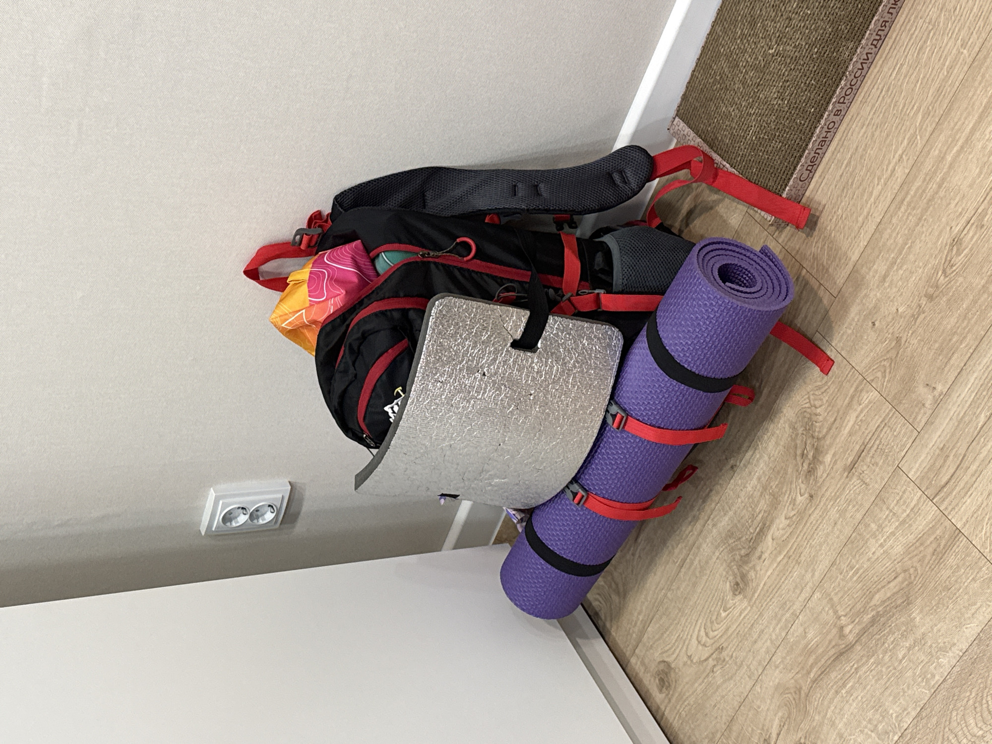 A backpack with a rolled purple mat and a reflective pad attached is on the floor. The location appears to be indoors, in Izhevsk, Russia based on the inscription on the edge of a carpet in the scene. The backpack is black with red straps and a partially visible multicolored bag. A dual electrical outlet is visible on the white ceiling. The floor has a light-colored wood-grain pattern.