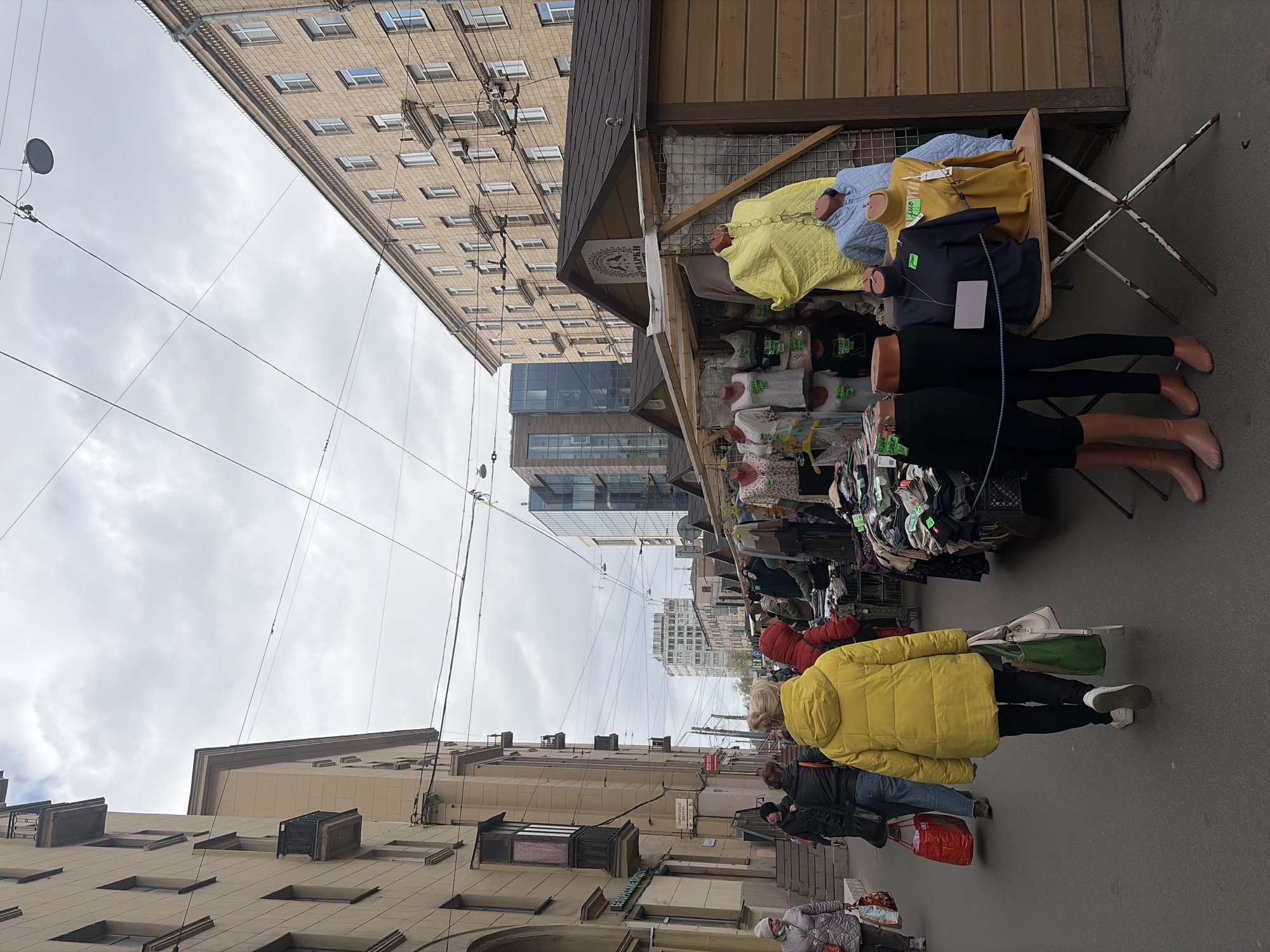 The image depicts a street scene in Saint Petersburg, Russia. A line of street vendors displaying clothing on mannequins occupies the right side of the frame. Customers, wearing warm winter clothing, are visible walking along the sidewalk next to the vendor stalls. Buildings with architectural details typical of the city line both sides of the street, and power lines stretch across the sky above. The weather appears overcast, with a cloudy sky.