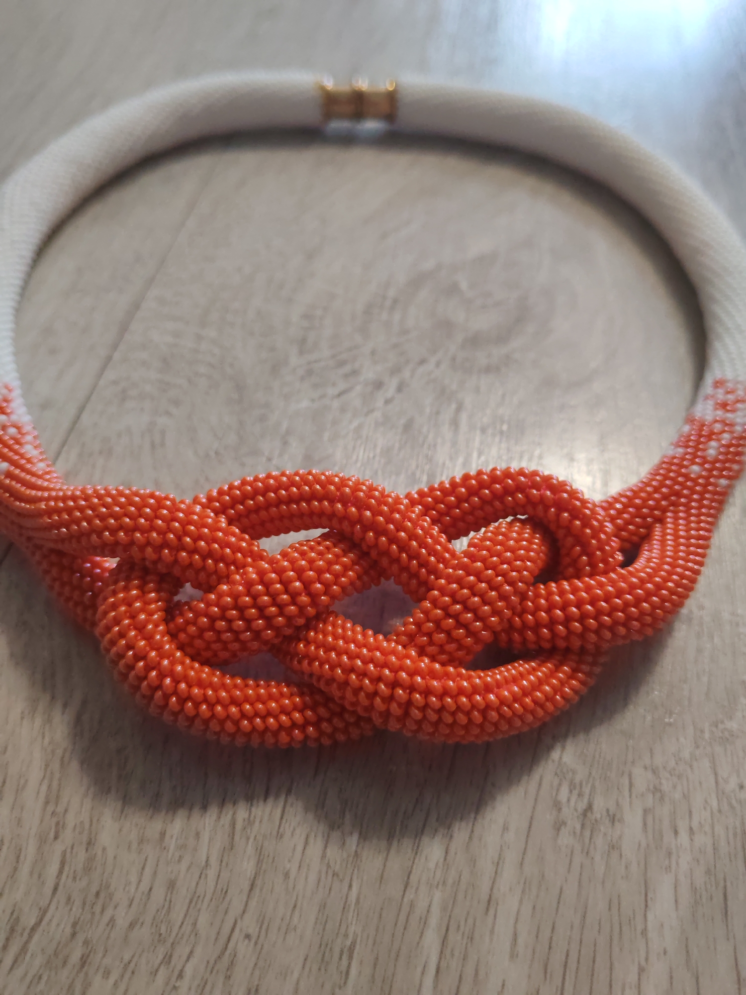 A beaded necklace is laid out on a wooden surface. The necklace is circular and made of small beads. The majority of the necklace is white, with a transition to orange beads in the front. The orange beads form a knot-like design. The necklace has a gold clasp. The wooden surface has a visible grain pattern.