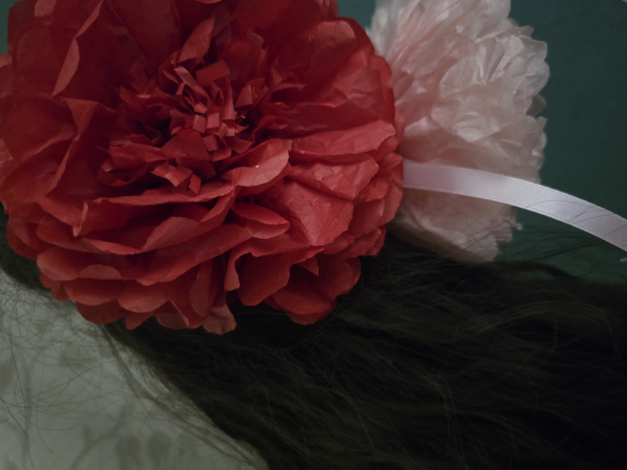 A close-up, vertical, low-angle shot shows a large, red, paper flower decoration. The flower is situated to the right of the frame, with its textured petals dominating the center. To the left, dark, wispy hair is visible, suggesting the flower is attached to someone's head. Below the red flower, a smaller, lighter pink, paper flower decoration is visible, connected to the red one by a white ribbon that hangs down the left side of the frame. The background is a mottled light gray on the left and a dark teal on the right. The scene appears to be indoors, likely a domestic setting.