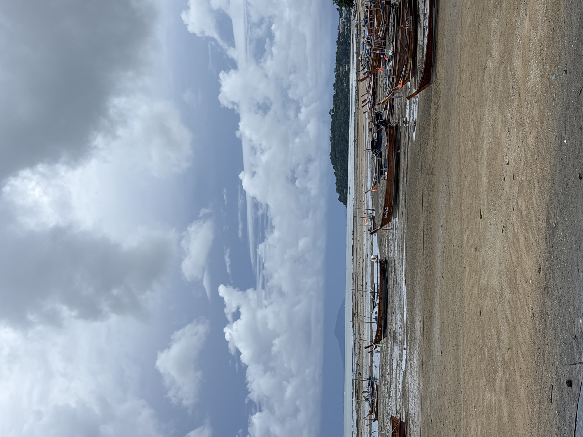 The image depicts a beach in Rawai, Thailand, characterized by a muddy shoreline with numerous long-tail boats grounded due to low tide. The sky is overcast with dramatic, textured clouds. In the distance, a lush green island is visible across the water, with some structures on its slopes. The foreground shows the sandy, wet beach, interspersed with shallow pools of water. The boats, made of wood with distinctive long poles at their sterns, are positioned in various orientations on the mud. Some boats have people inside or near them, though their activities are not clearly discernible. The overall atmosphere is somber, likely due to the cloudy weather.