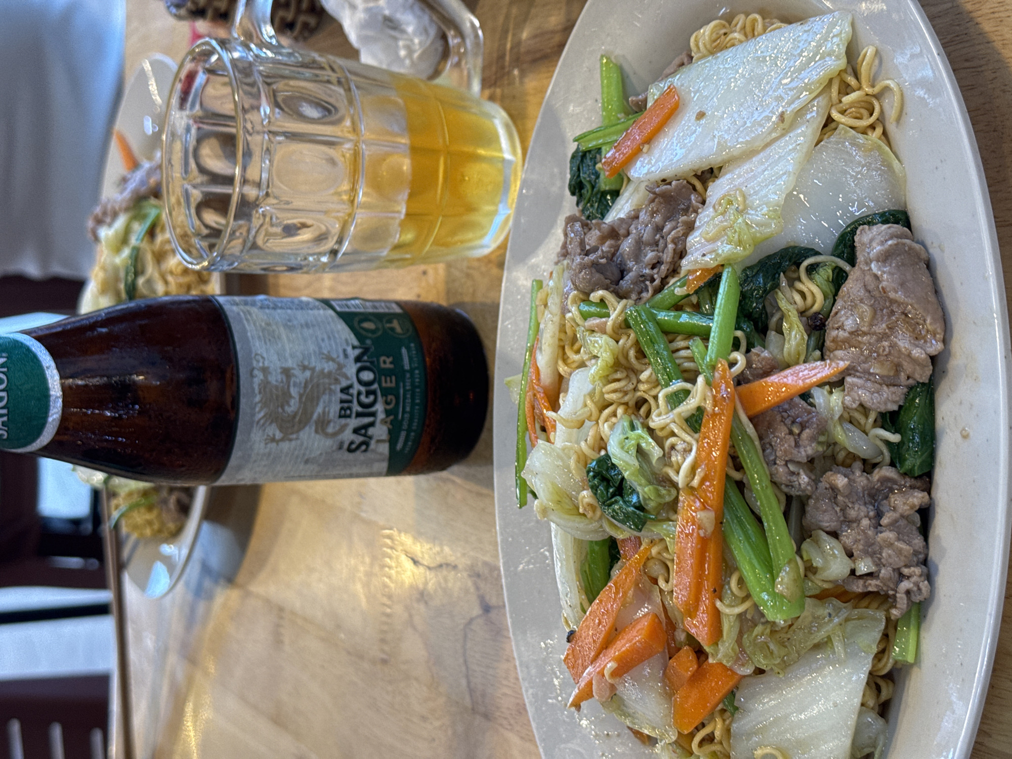 On a wooden table, a half-full mug of beer sits beside a bottle of Bia Saigon Lager. A plate of stir-fried noodles with meat and vegetables is arranged to the right of the beverages. Another plate with food is partially visible in the background. The setting is an unidentified location within an unknown city.
