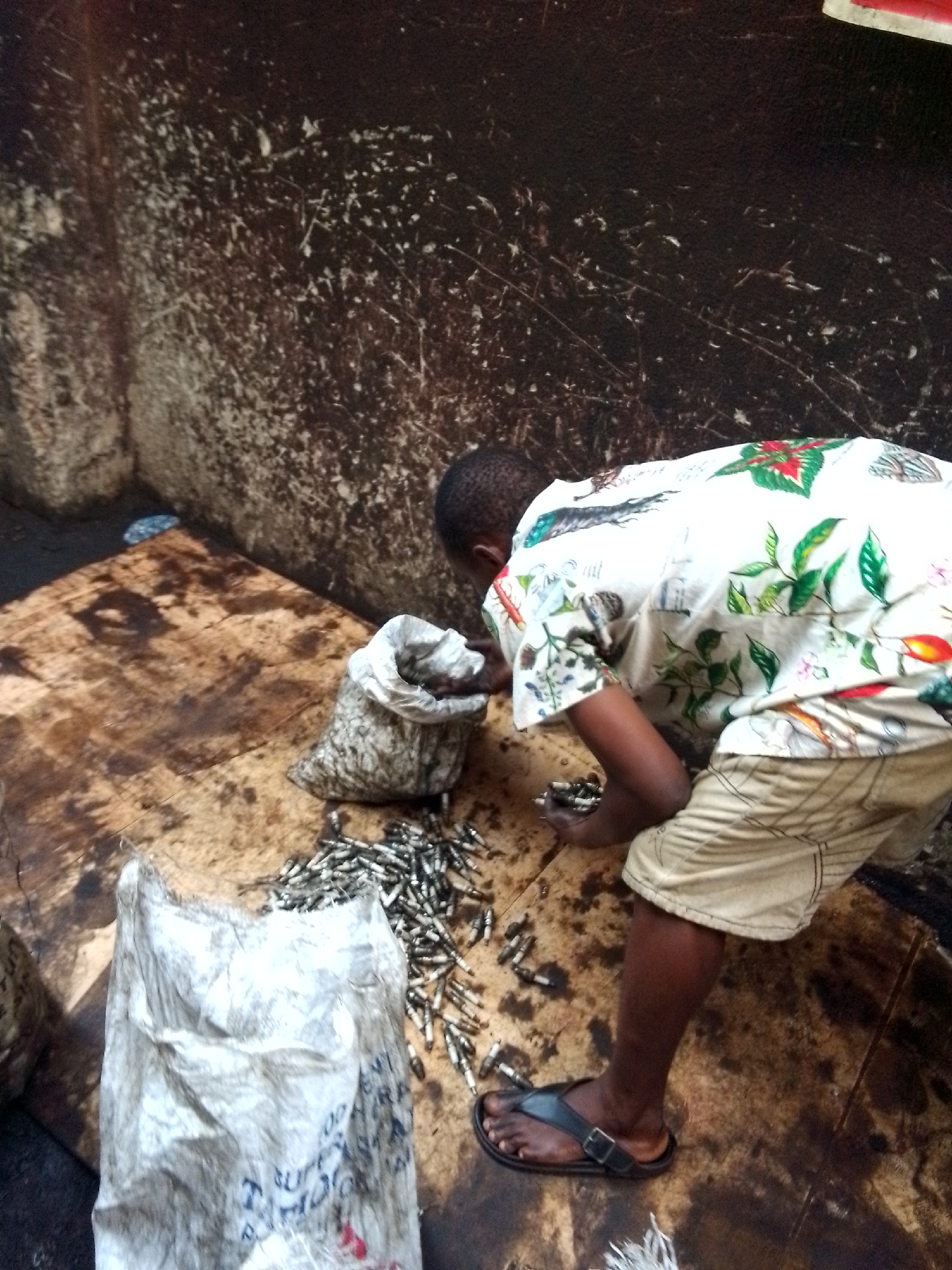 A man in Onitsha, Nigeria, sorts spark plugs at an outdoor workstation. The man wears a patterned white shirt, khaki shorts, and black sandals. He stands on a stained floor in front of a stained wall. A worn wooden table holds two bags of spark plugs and a pile of loose spark plugs, which he is sorting.
