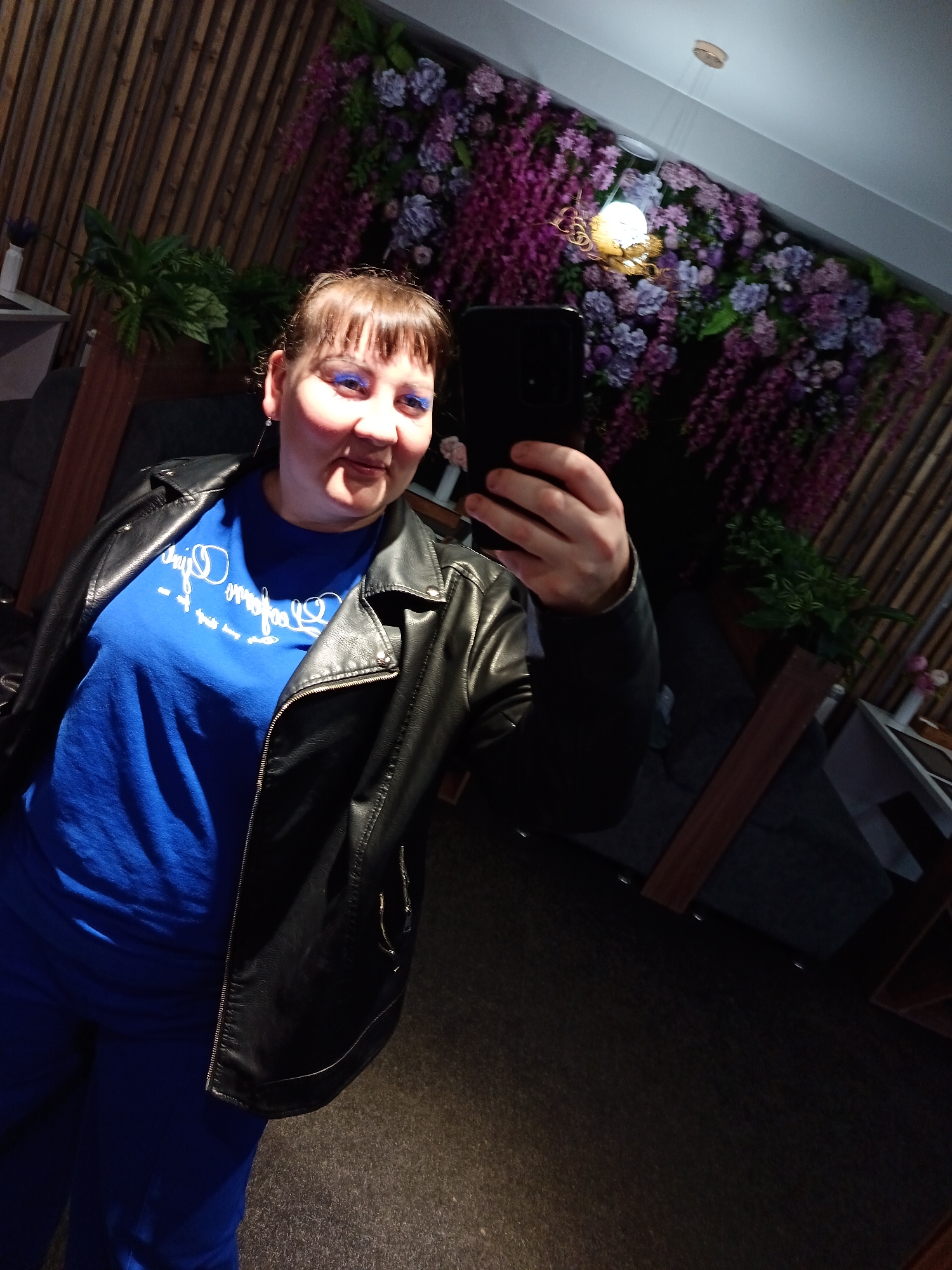 An indoor selfie captures a smiling woman in Osa, Russia, with a cheerful expression and bright blue eyeshadow. She has light brown hair with bangs and is holding a dark smartphone to take the picture. She is wearing a vibrant blue t-shirt, a black leather jacket with zippers, and matching blue trousers. White, stylized cursive text is visible on her t-shirt, appearing mirrored due to the selfie angle, and is difficult to decipher definitively, but seems to contain words like "Dream."
The background features a decorative wall. To the left, there are vertical wooden slats, while the right side is adorned with a large arrangement of artificial purple and pink flowers or vines, creating a lush, floral display. A decorative light fixture with a glowing bulb hangs in front of the floral wall. Green potted plants are also incorporated into the decor. Dark furniture, possibly sofas or chairs, and tables are partially visible in the dimly lit setting. The floor is dark and textured. The scene suggests an evening or nighttime atmosphere in an interior space, such as a restaurant or lounge.
