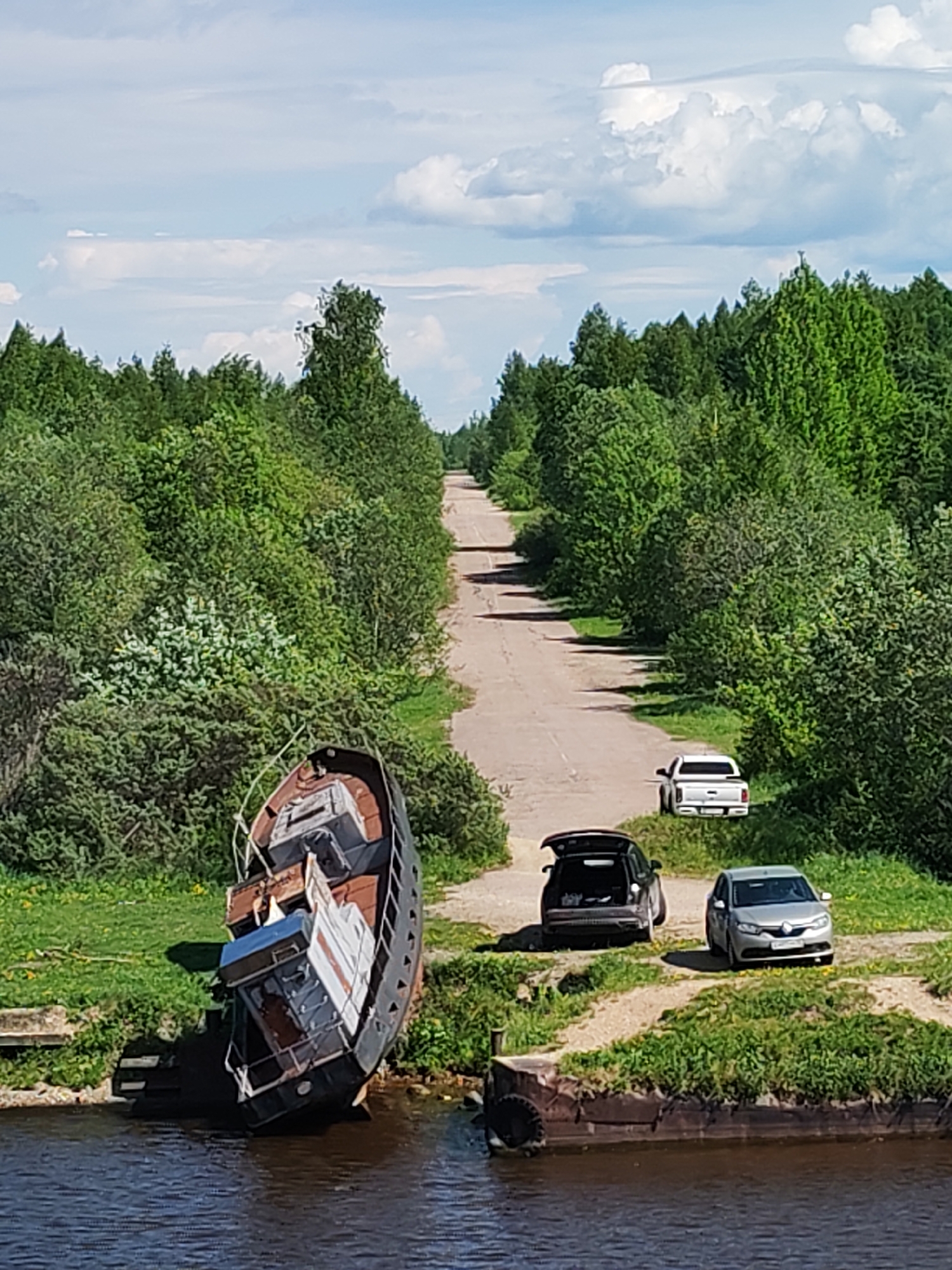 The image shows a landscape in Ivanov Bor, Russia, featuring a road leading into a forest under a cloudy sky. A derelict boat is partially submerged in water in the foreground. Three vehicles are parked alongside the road: a silver sedan, a dark SUV with its hatch open, and a white pickup truck further down the road. The environment includes lush green trees lining the road and bodies of water surrounding the boat and pier.