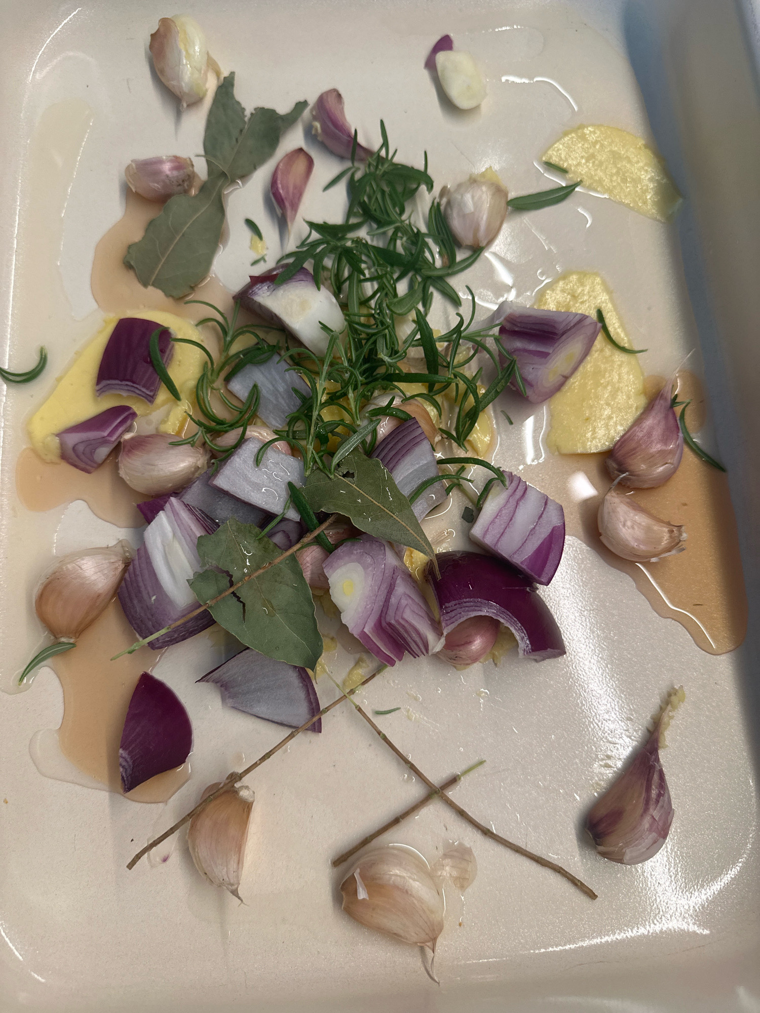 A close-up, overhead shot shows a collection of ingredients in a light-colored rectangular baking dish, possibly preparing for cooking. The dish contains several whole and separated garlic cloves, some appearing peeled and others with their skins intact, some stained purple. Chunks of red onion, cut into wedges, are scattered throughout the dish, revealing their layered purple and white flesh. There are also several dried bay leaves, a sprig of fresh rosemary, and thin slices of what appears to be ginger or perhaps a yellow root vegetable. The ingredients are resting in a light-colored liquid, likely oil or rendered fat, which pools in some areas of the dish. The background is the white ceramic or enamel interior of the baking dish. There are no people or animals visible in the image. The scene does not provide any information to indicate a location in Nevinnomyssk, Russia.