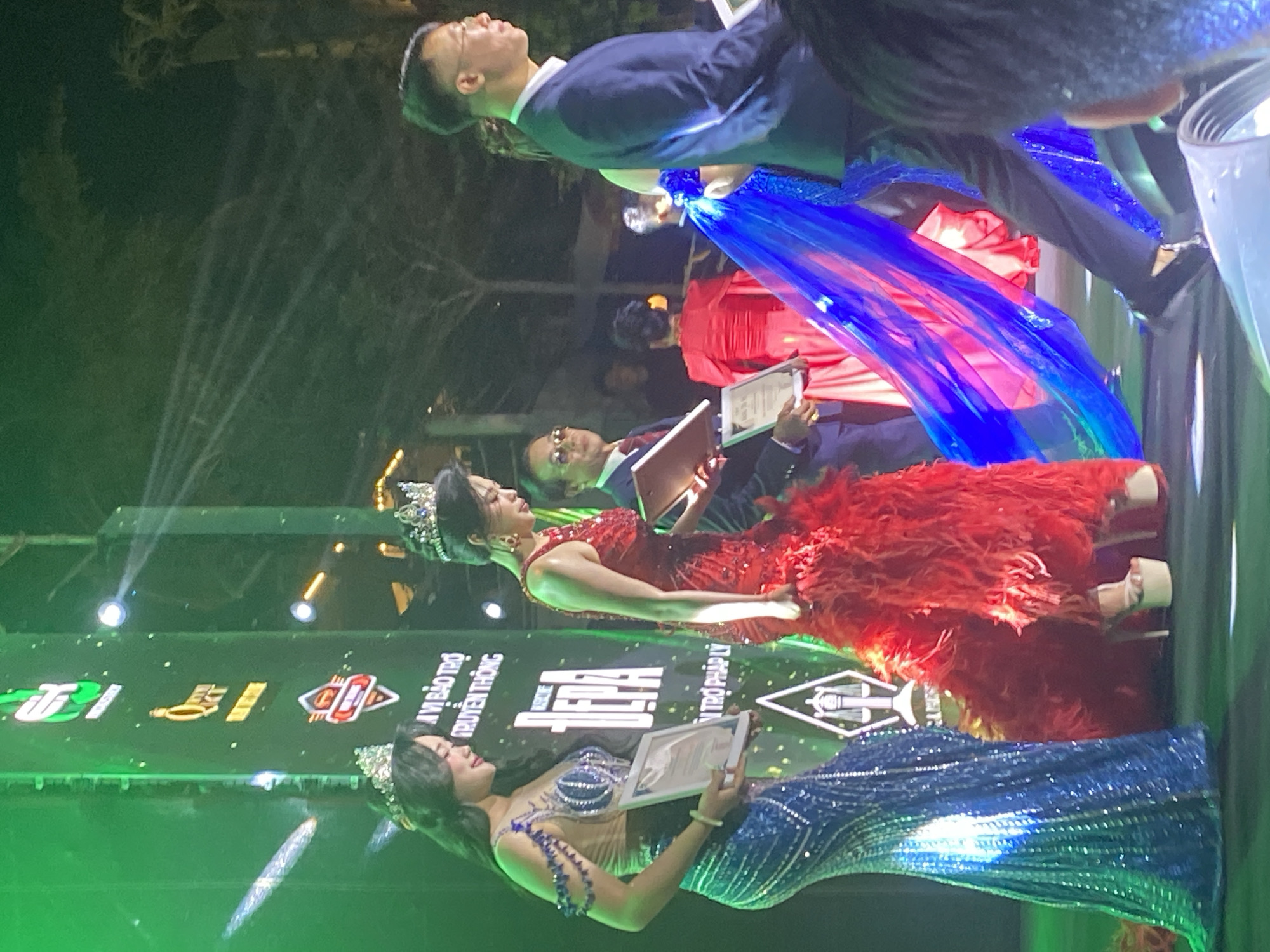 In Mũi Né, Vietnam, a formal awards ceremony is underway on a stage illuminated by green and white lights. Two women, adorned with sparkling crowns and elaborate gowns, stand on the stage. The woman on the left wears a shimmering blue dress with beaded embellishments and holds a framed certificate. The woman to her right is dressed in a vibrant red gown with a feathered hem and also holds a certificate. Several individuals in formal attire, including men in suits and women in formal dresses, are present on the stage, some of whom appear to be presenting awards. A large LED screen in the background displays event logos and text. The atmosphere suggests a prestigious event, likely celebrating achievements in a particular field, with the backdrop of Mũi Né's coastal setting implied by the event's location.